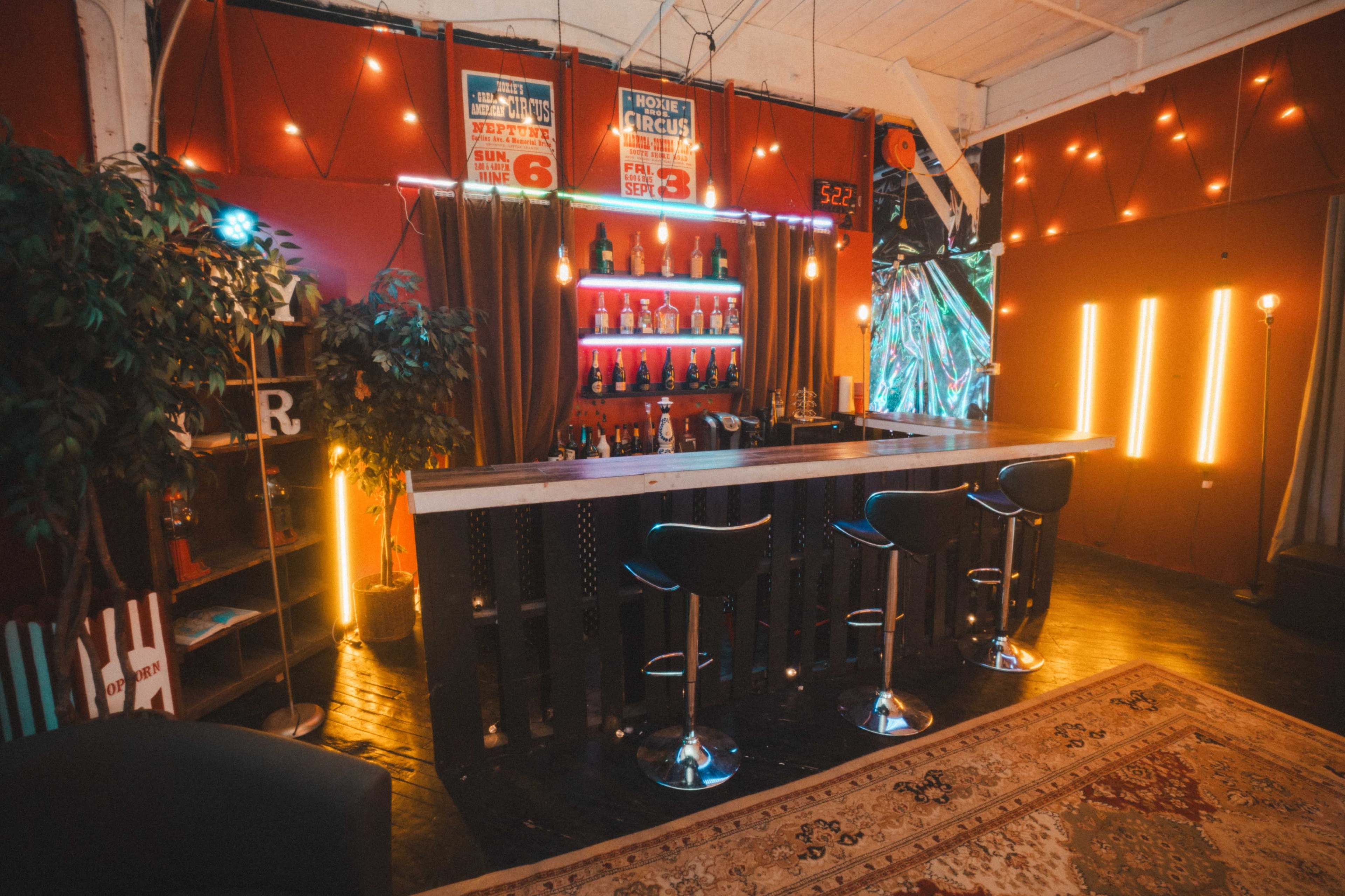 The image shows a well-lit bar area with a wooden countertop, high stools, illuminated bottles on shelves, and colorful neon lights along the wall.
