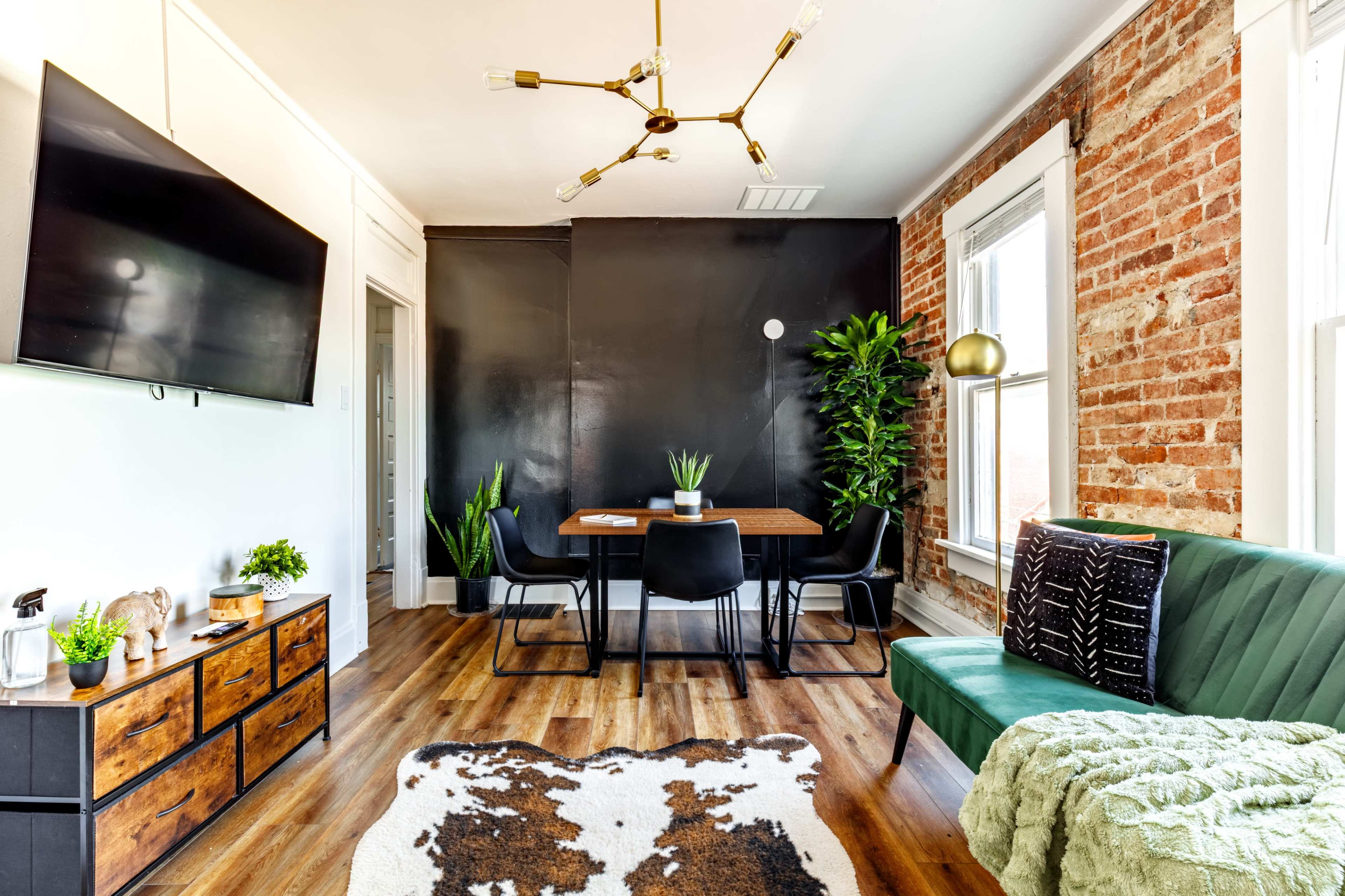 A modern living space features a black accent wall, a wooden dining table with chairs, and a combination of plant decor and various furniture pieces, including a green sofa and a television.