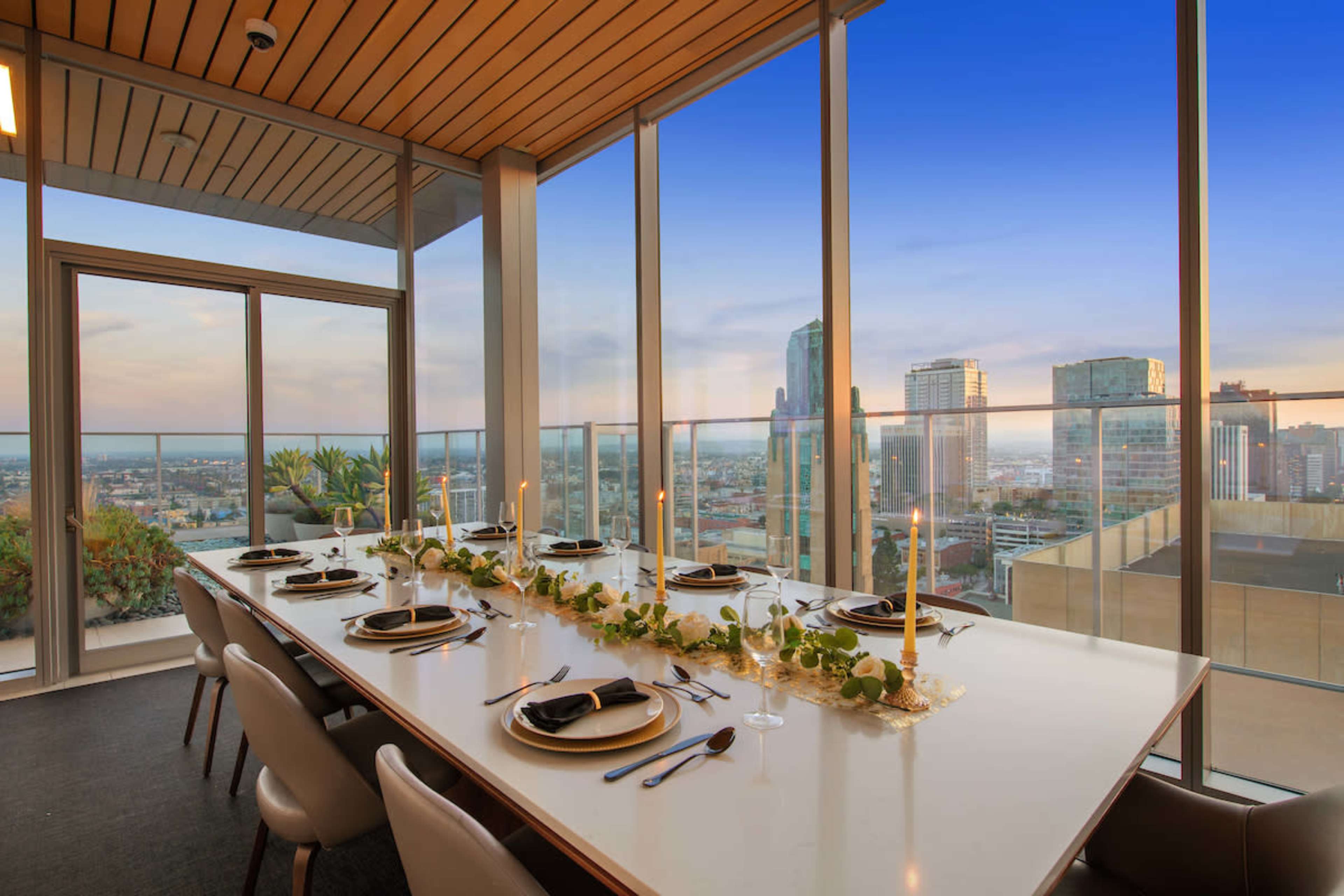 A long dining table is elegantly set with plates and candles, positioned by large windows overlooking a city skyline at sunset.
