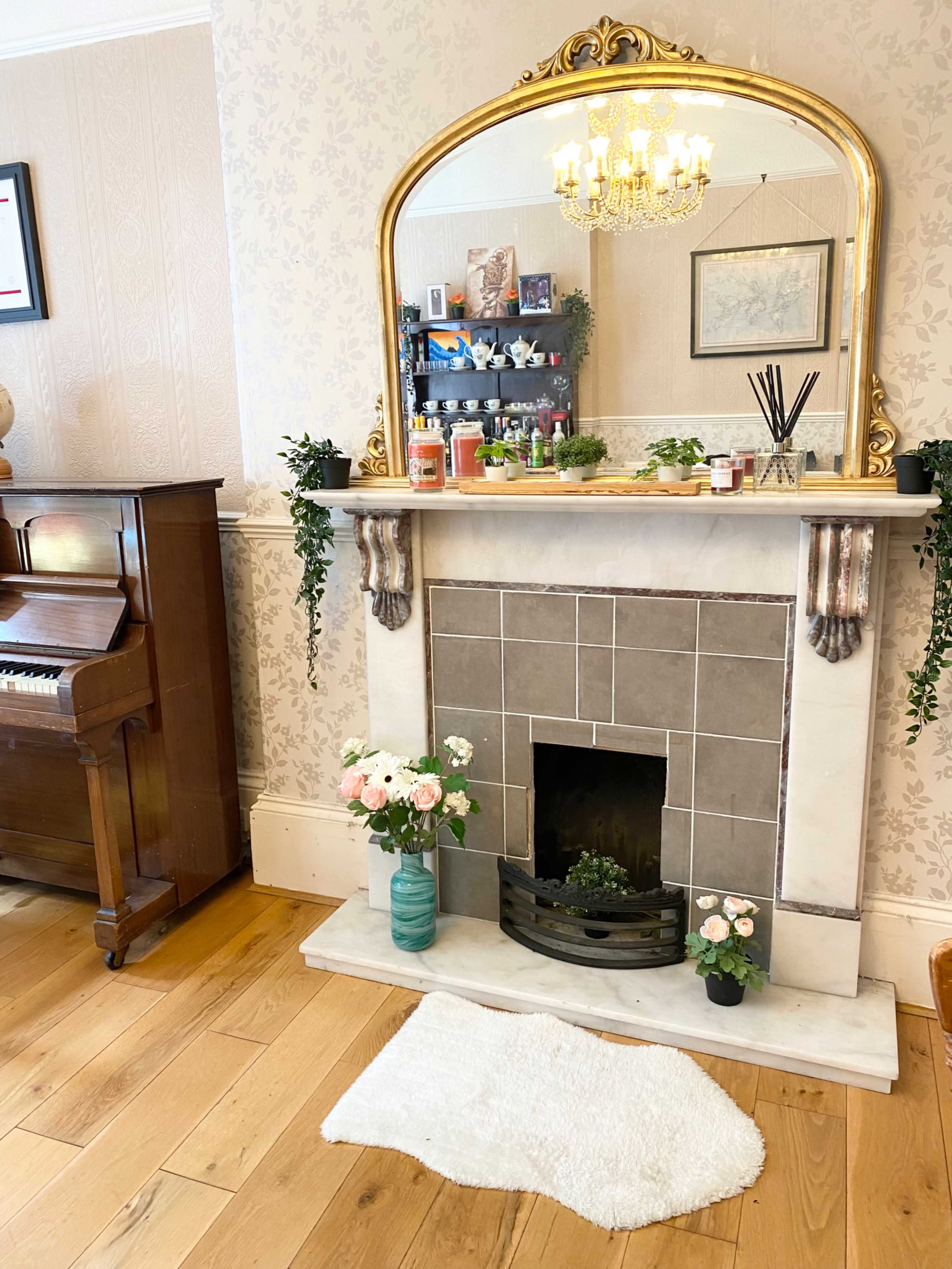 A decorative fireplace with a marble surround, topped by a large mirror and adorned with plants and candles, is situated next to a wooden piano on a hardwood floor.