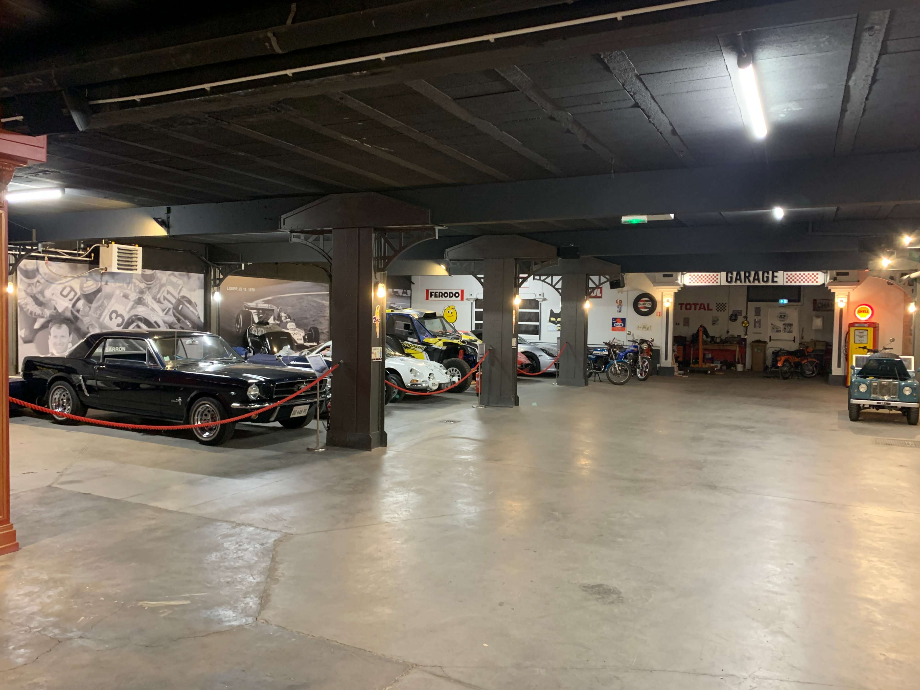 The image shows a spacious garage featuring several classic cars and motorcycles, with vintage signage and photographs on the walls.