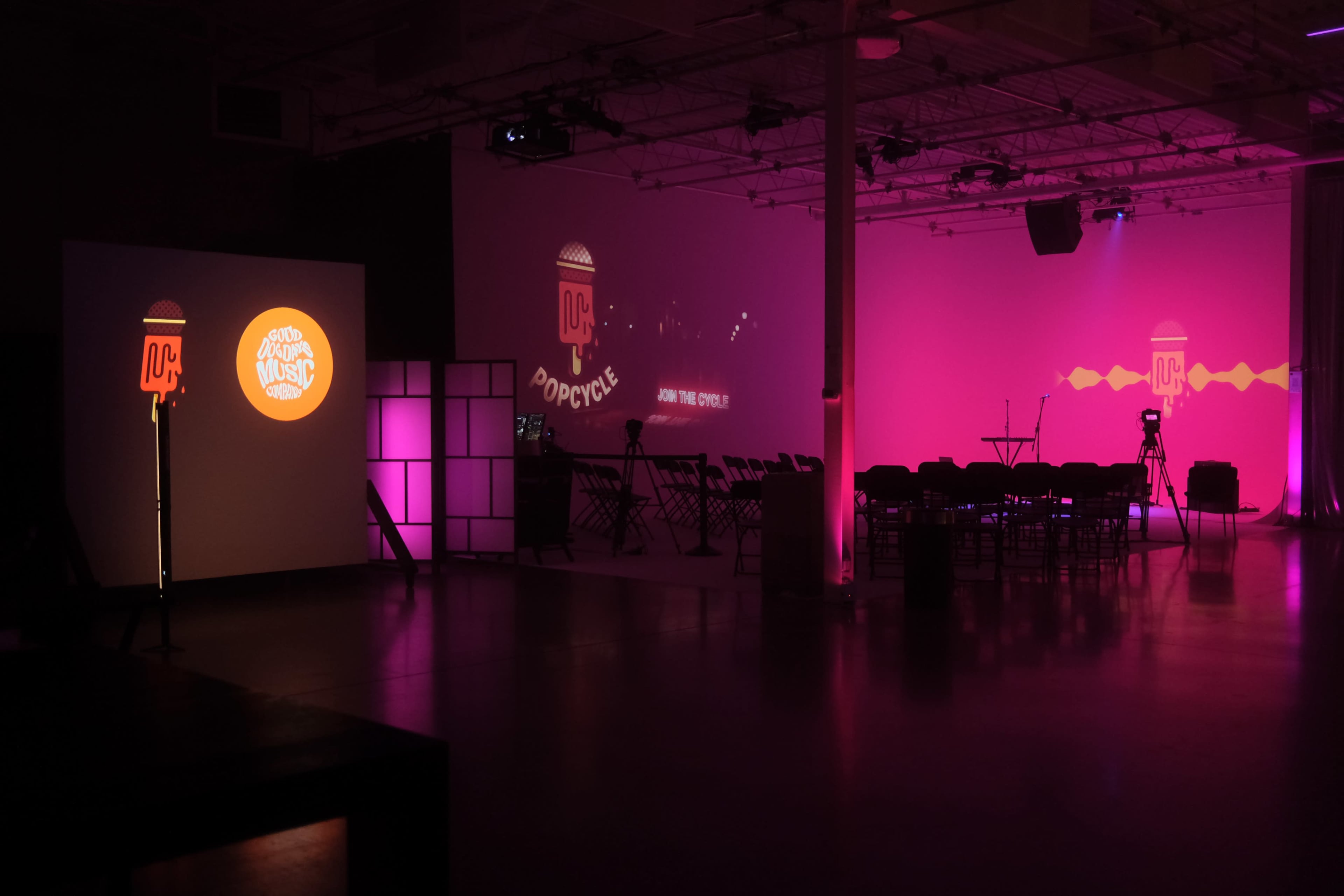 The image shows a spacious event venue with purple lighting, featuring a projection on a wall and seating arranged for an audience.