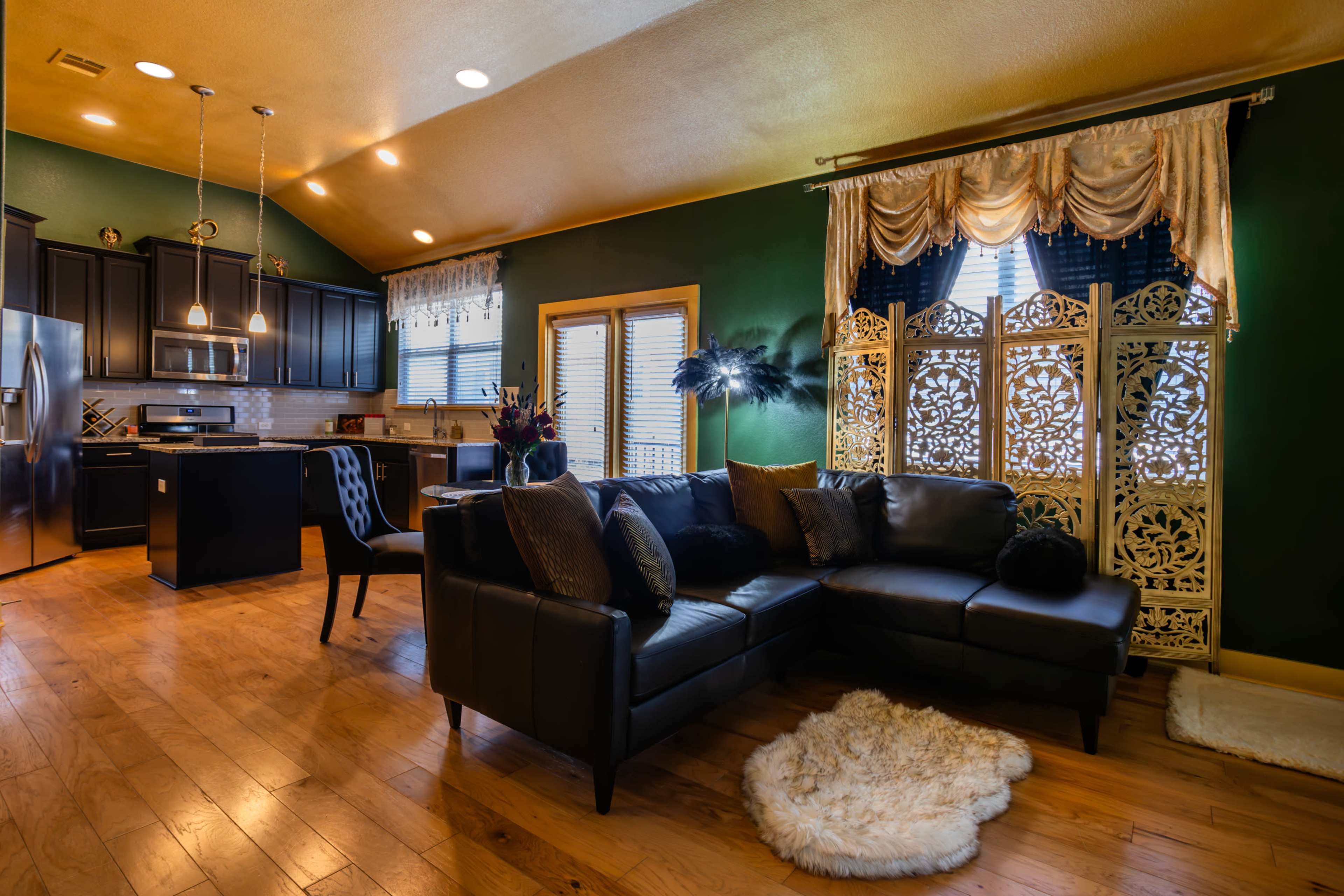 A cozy living room with a dark leather sectional sofa, wooden flooring, and a kitchen area in the background featuring black cabinets and stainless steel appliances.