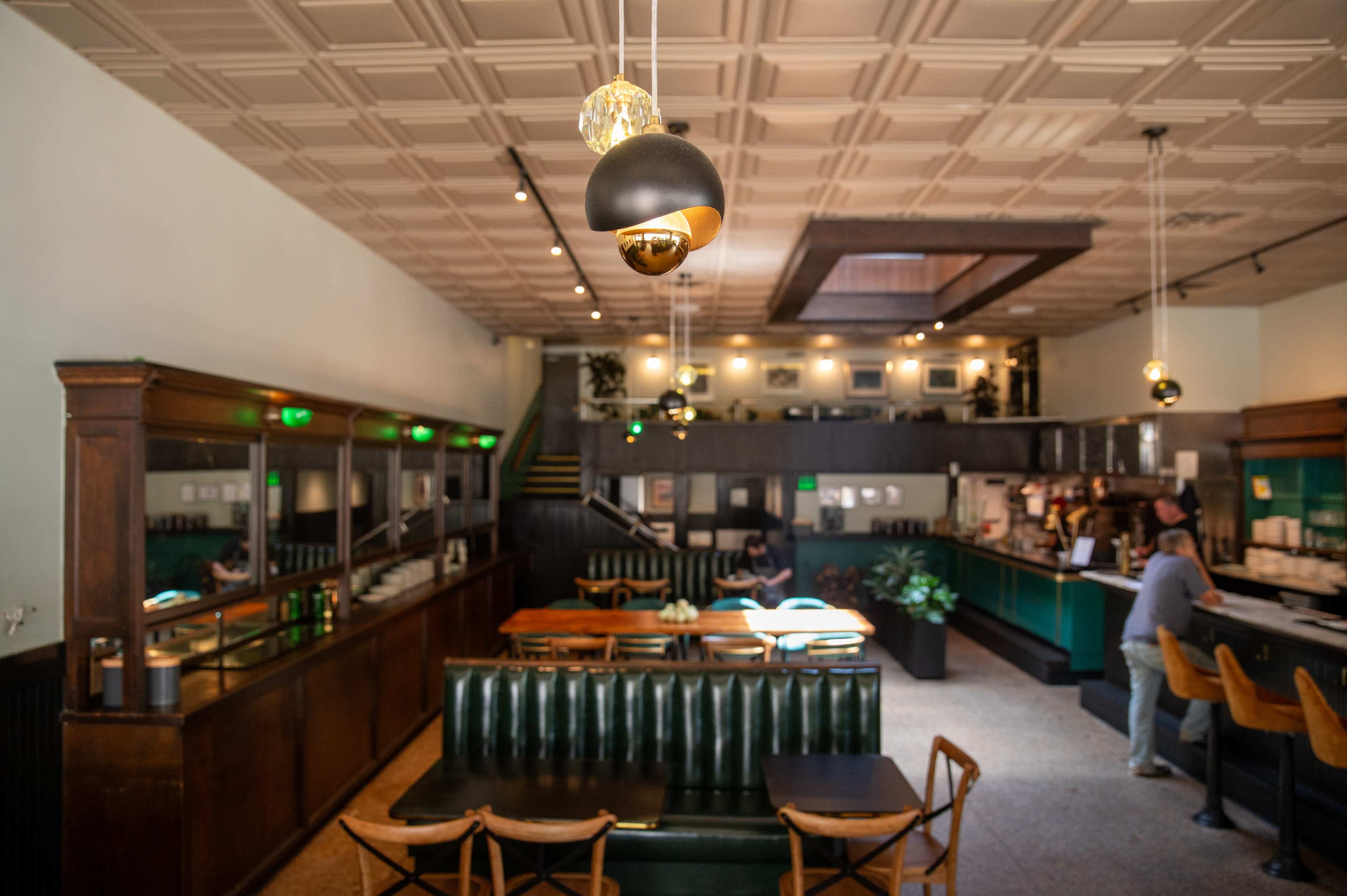The image shows a modern café interior featuring a mix of wooden and green leather seating, pendant lighting, and a coffee counter at the back.