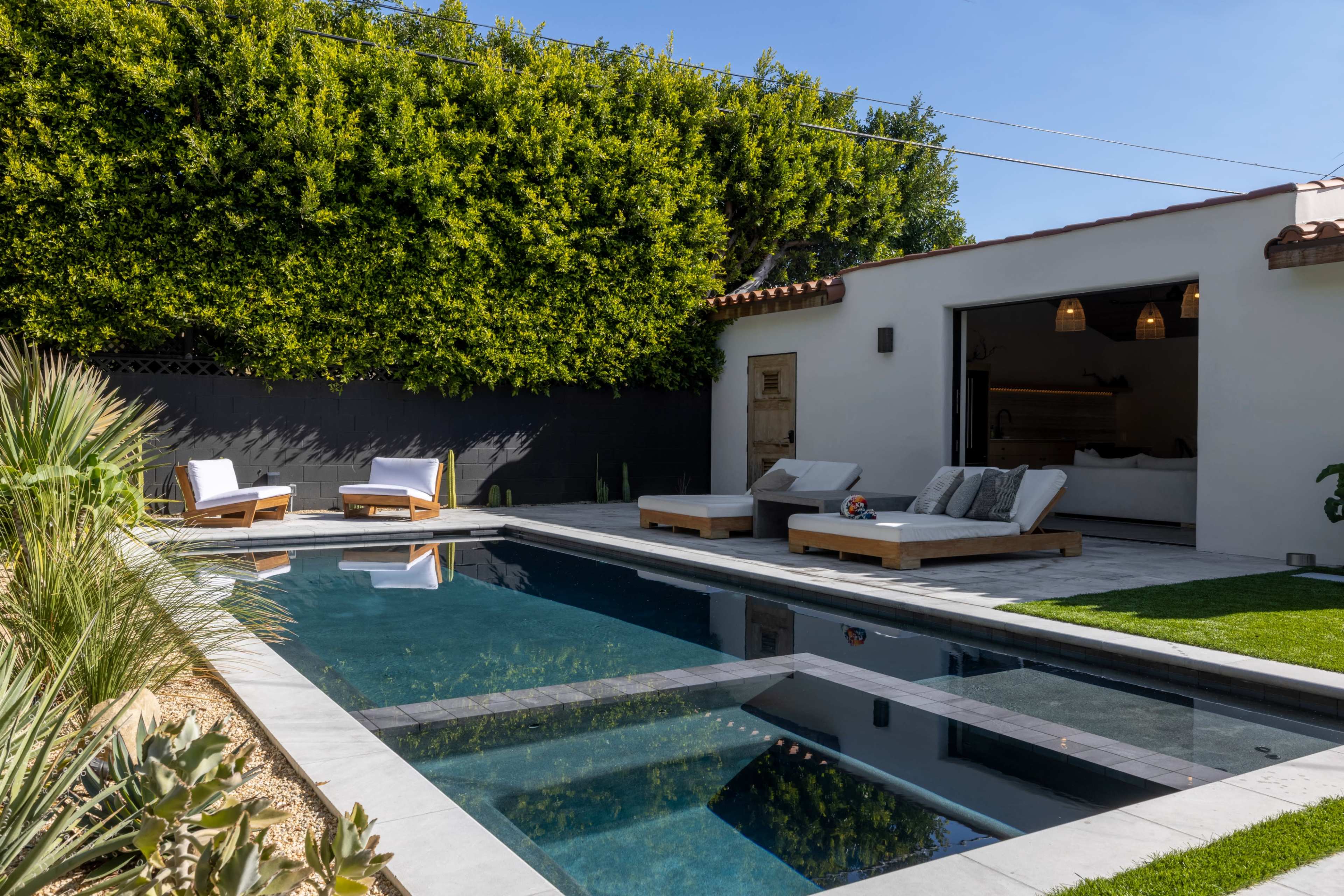 A modern backyard features a rectangular swimming pool surrounded by lounge chairs and landscaped greenery.
