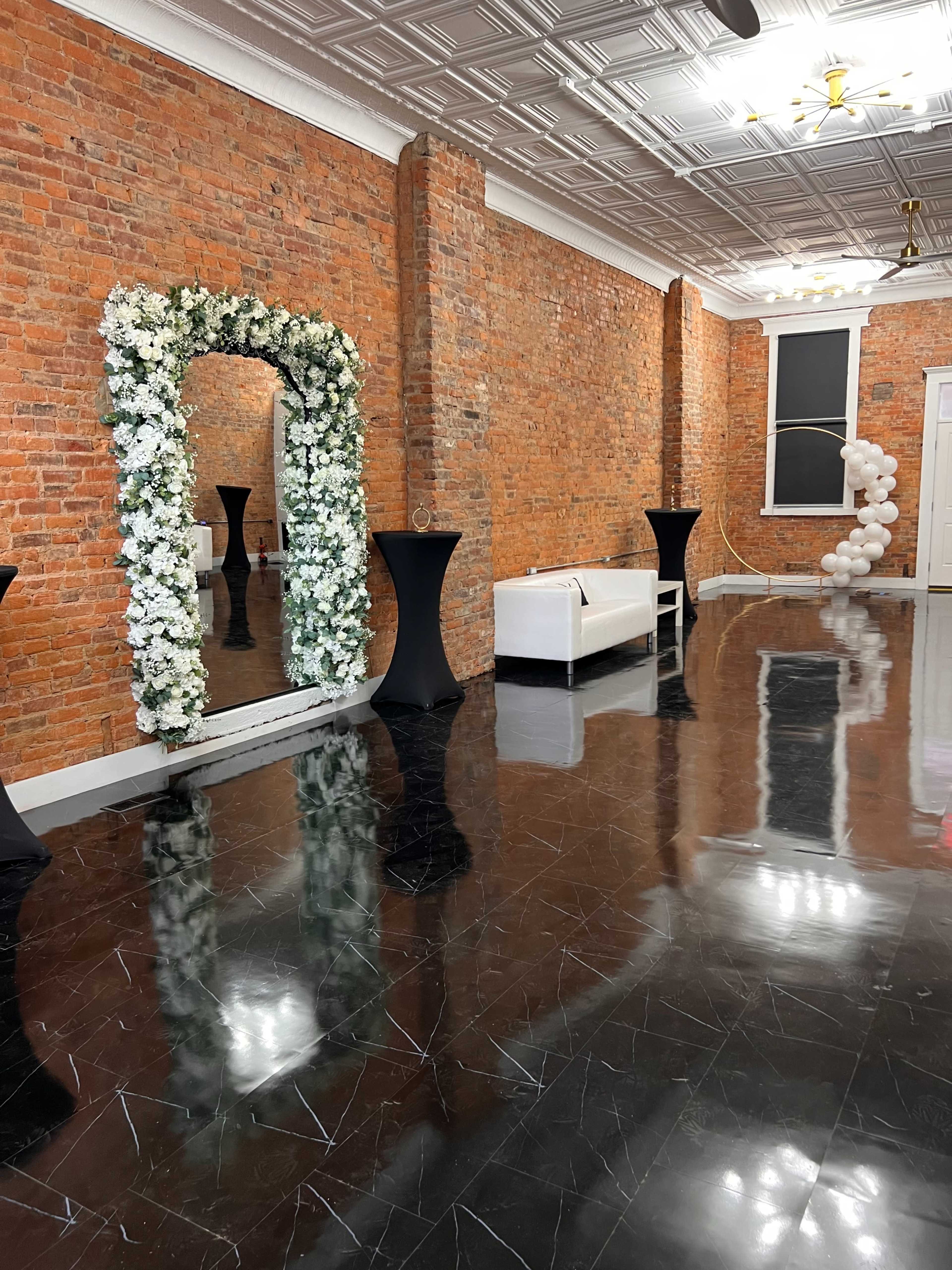 The image shows an empty interior space with exposed brick walls, a mirrored floral arch, and modern black and white furnishings.
