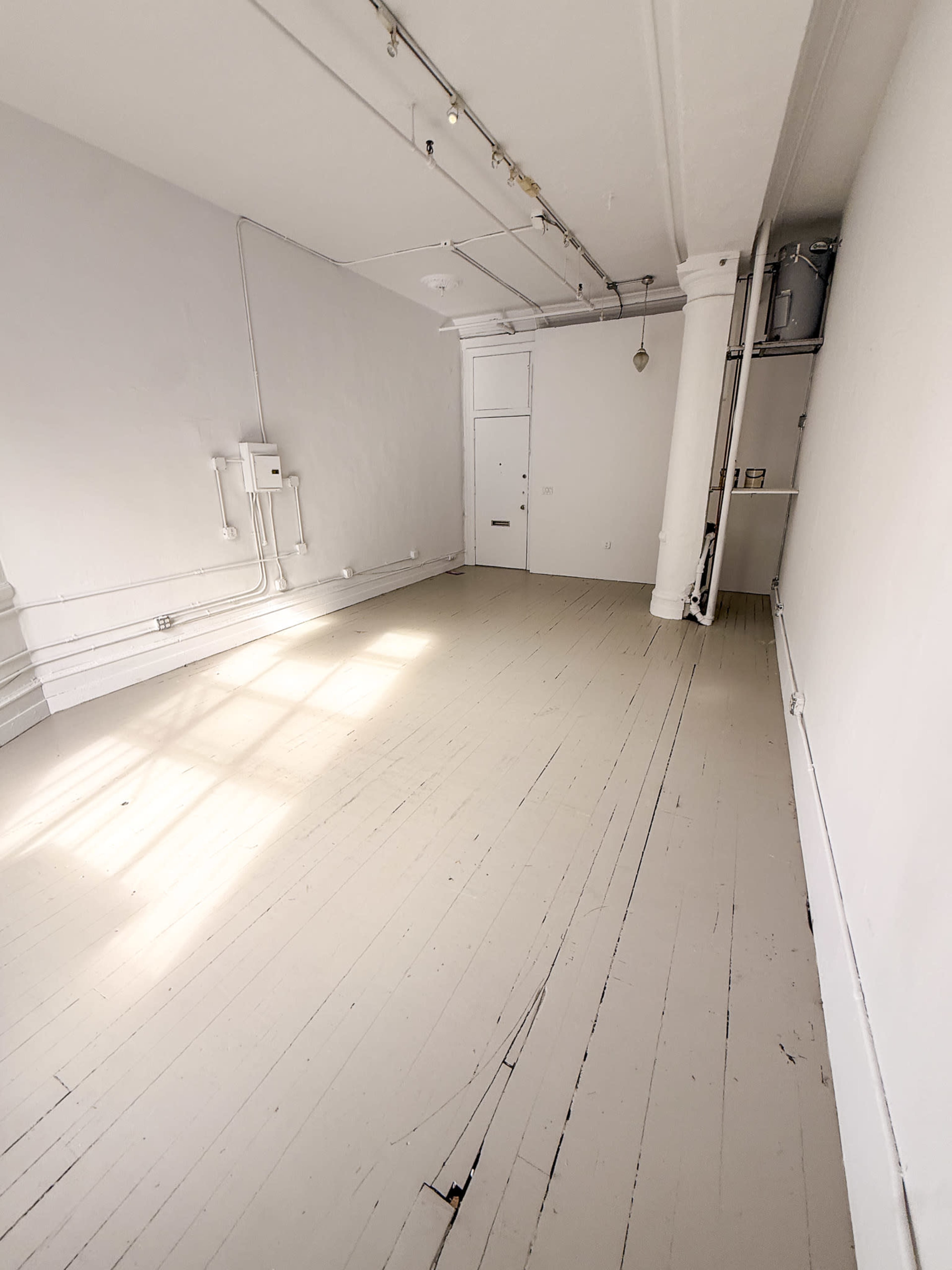 The image shows an empty room with light-colored wooden floors and white walls, featuring a single door and visible electrical fixtures.