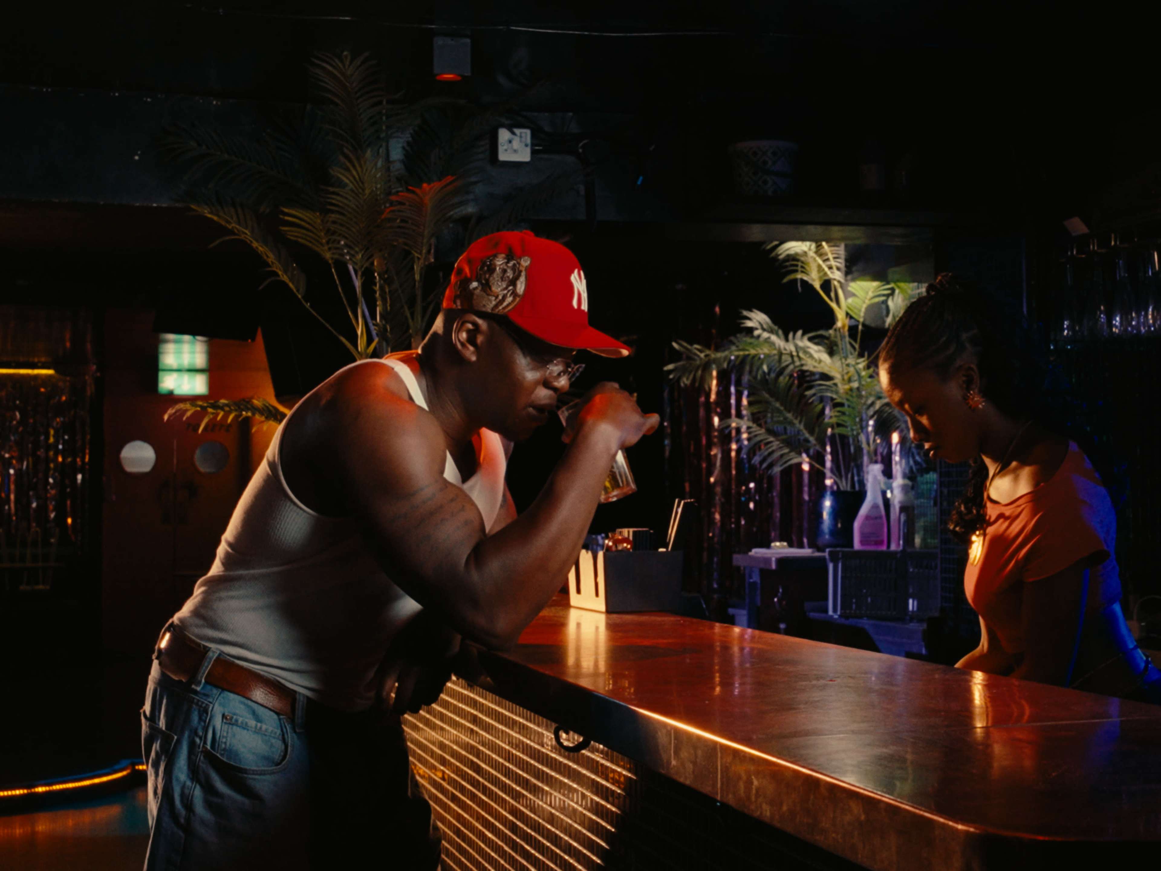 A man in a tank top and baseball cap leans on a bar while a woman stands behind it.
