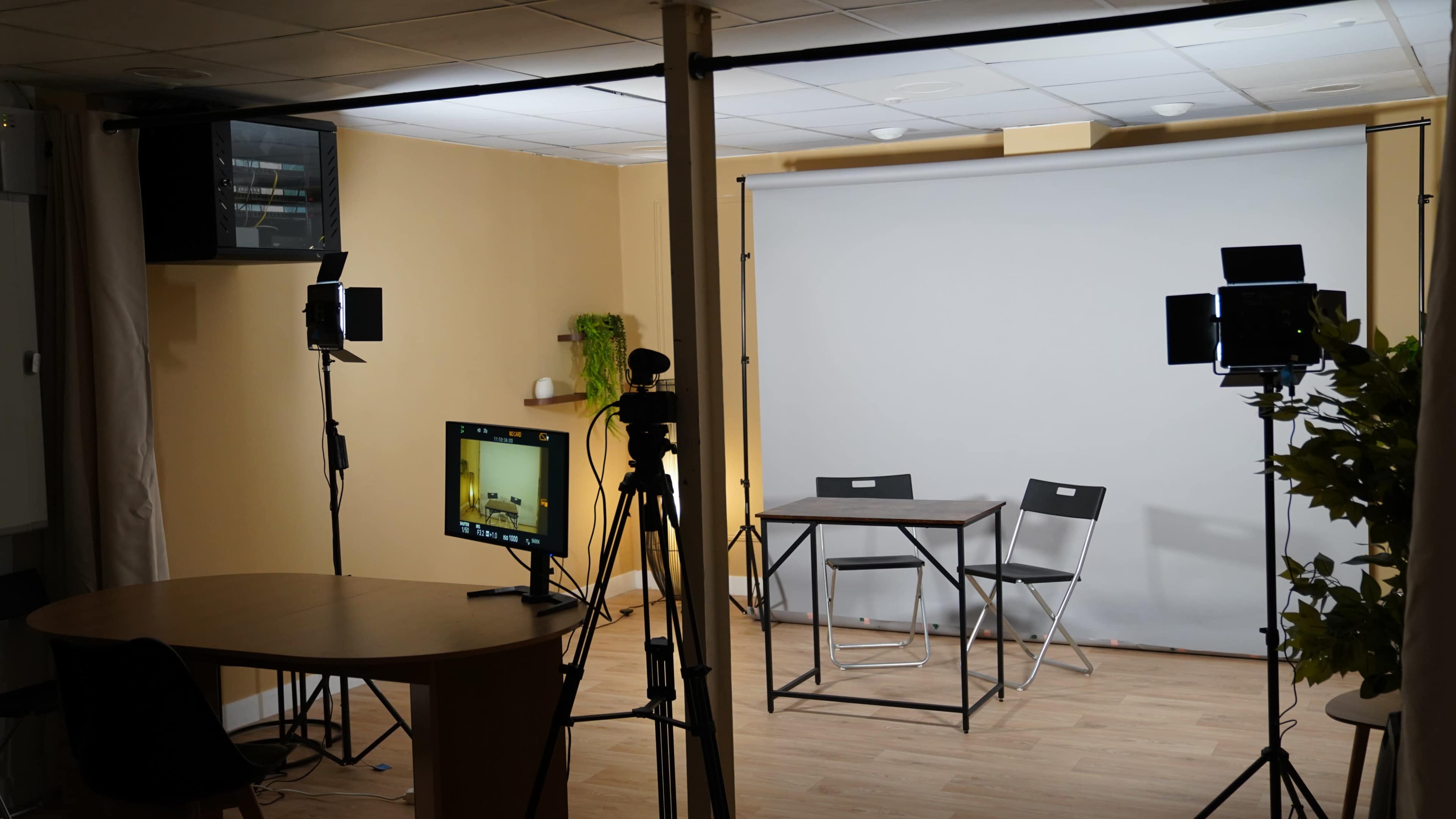 The image shows a small studio setup with a gray backdrop, two chairs at a metal table, and lighting equipment positioned around the space.