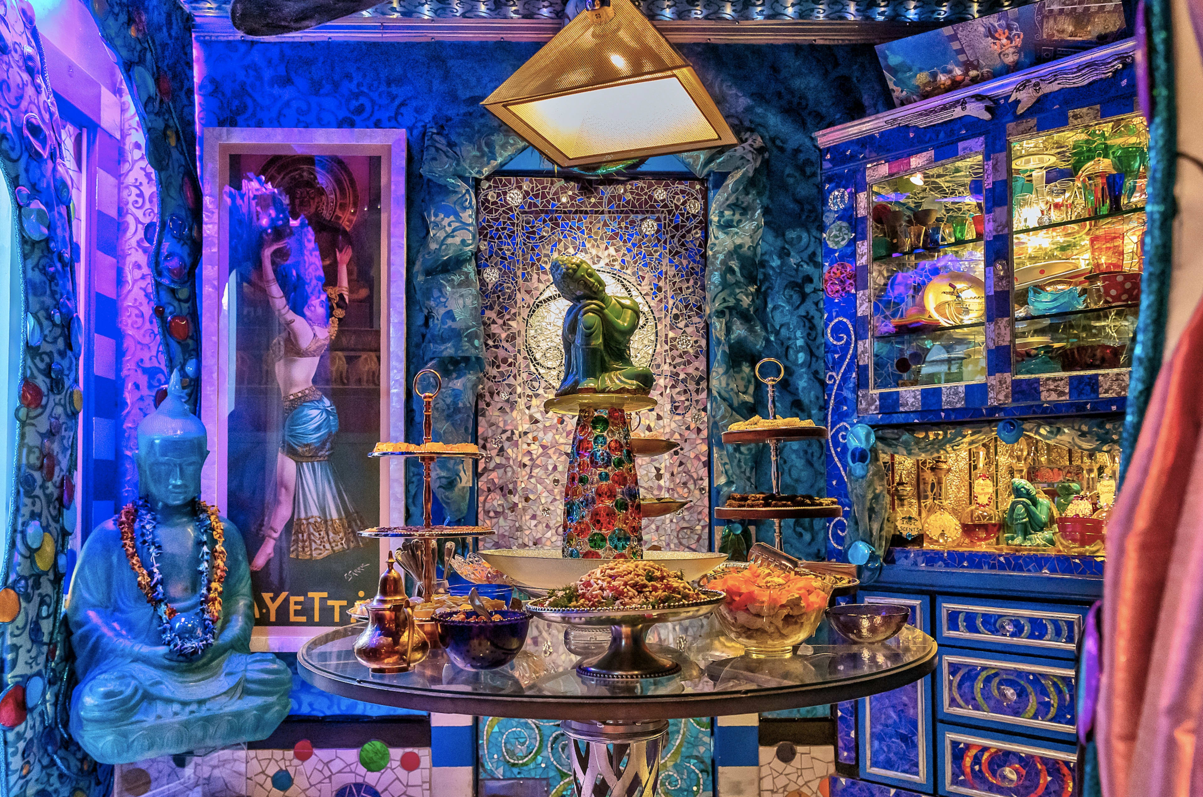 A vibrant, elaborately decorated room featuring a central table with a variety of dishes, flanked by intricate wall decorations and a statue of a seated figure.