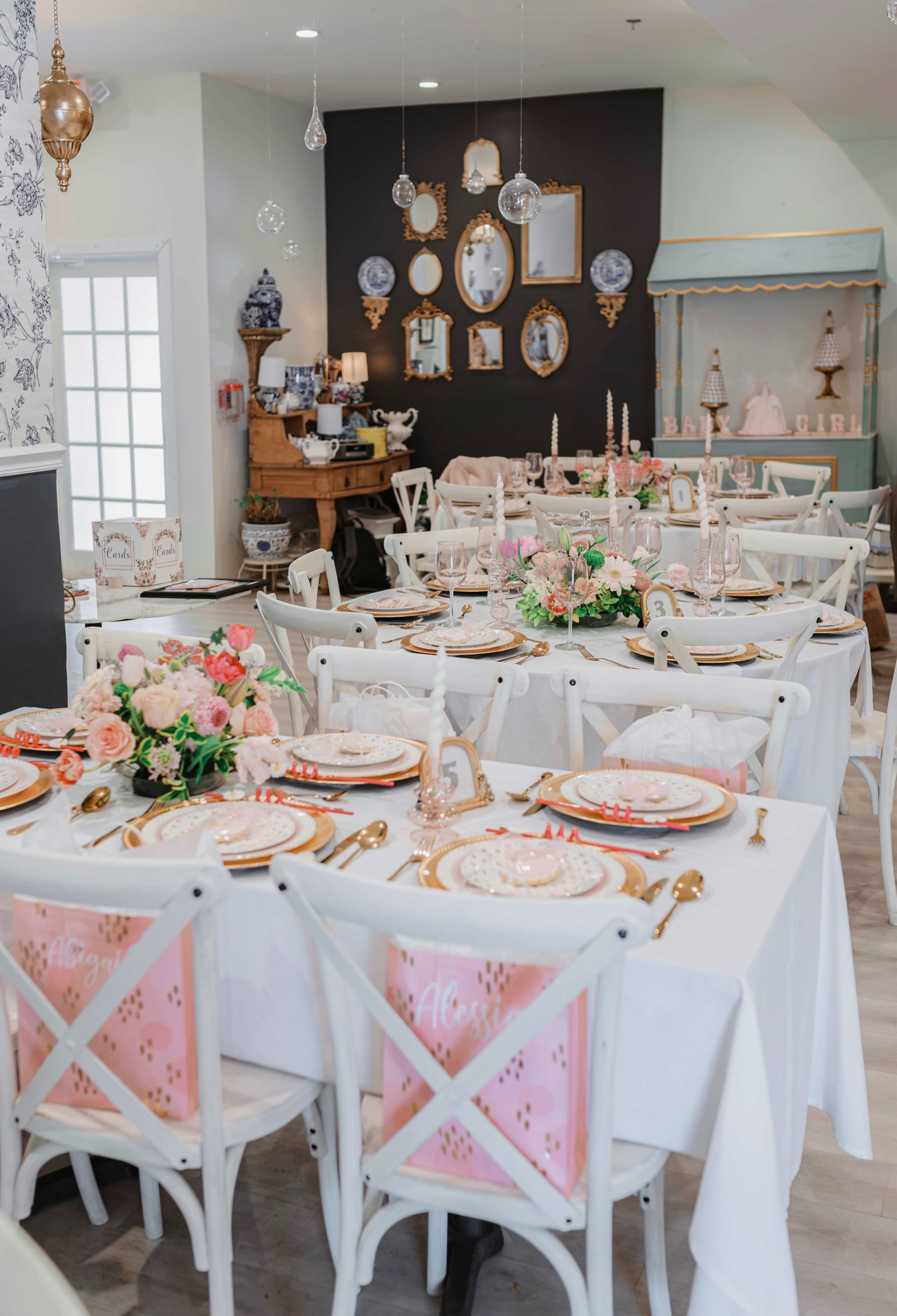A dining area is set with white tables draped in tablecloths, adorned with pink accents, floral centerpieces, and decorative plates, surrounded by white chairs and framed mirrors on the walls.