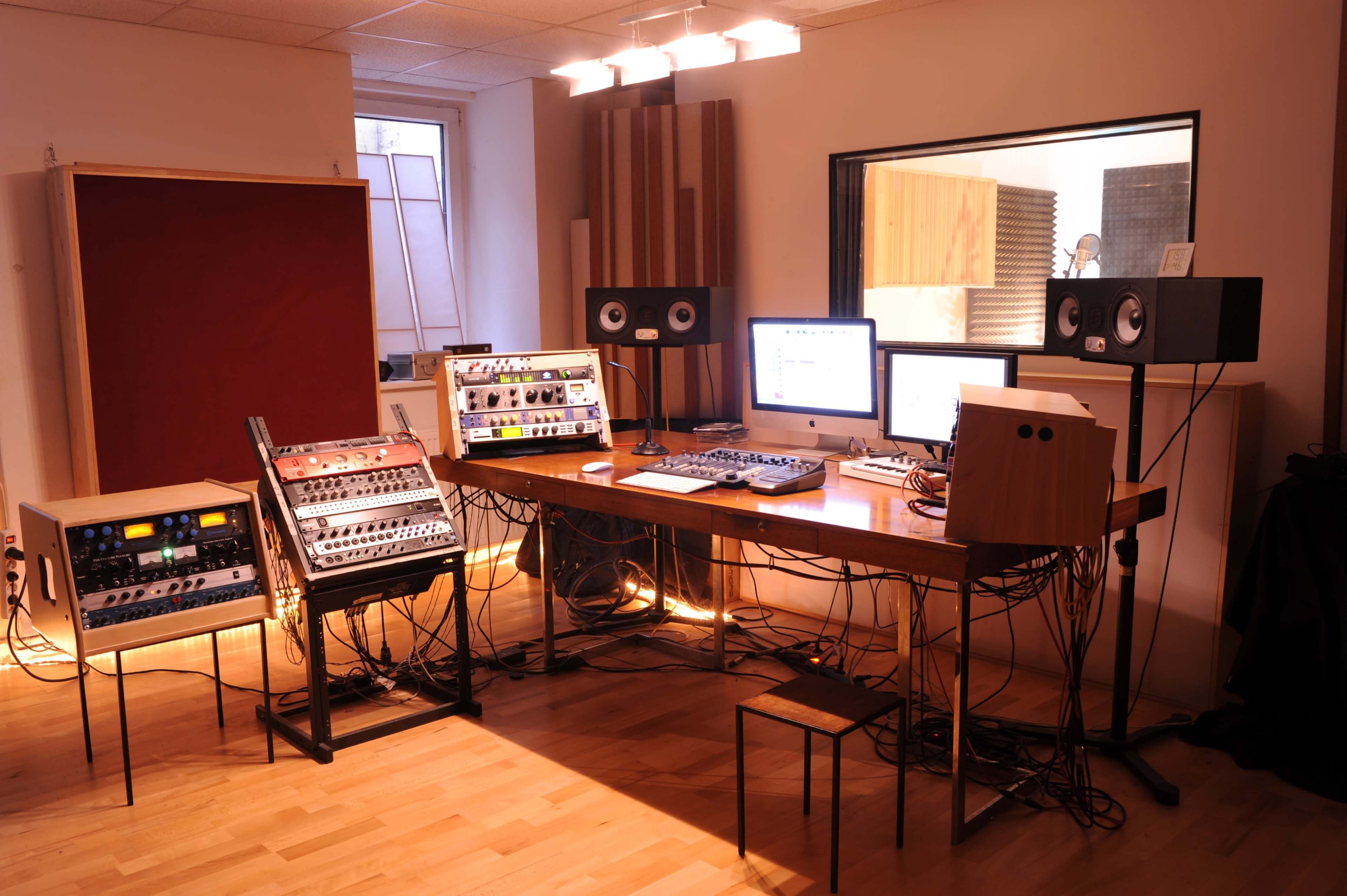 A music studio with audio equipment, a mixing desk, and monitors arranged around a wooden table in a well-lit room.