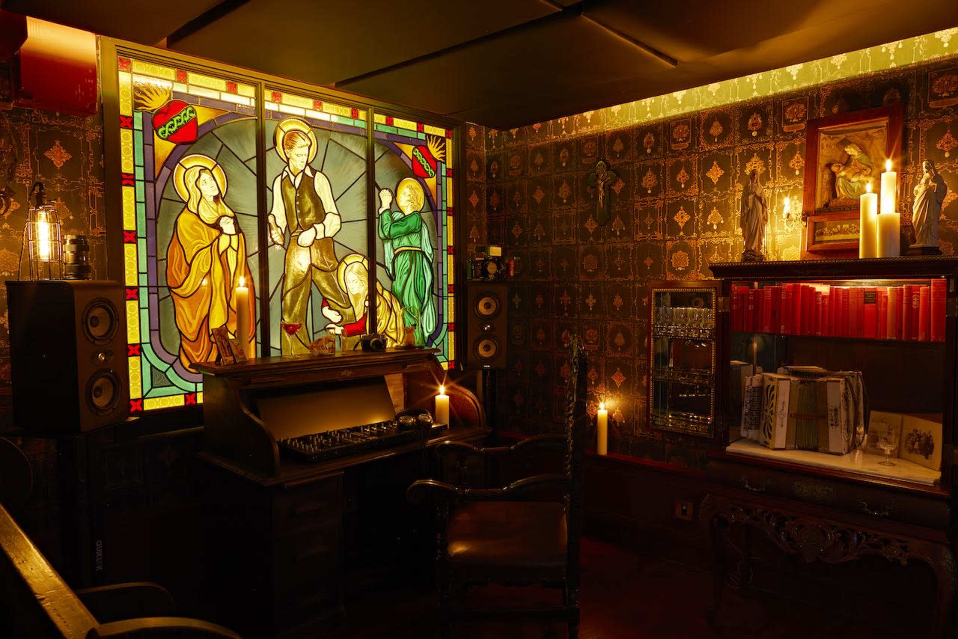 A dimly lit room with a stained glass window depicting three figures, an antique wooden desk with an old typewriter, a bookshelf filled with books, and various candles providing ambient light.