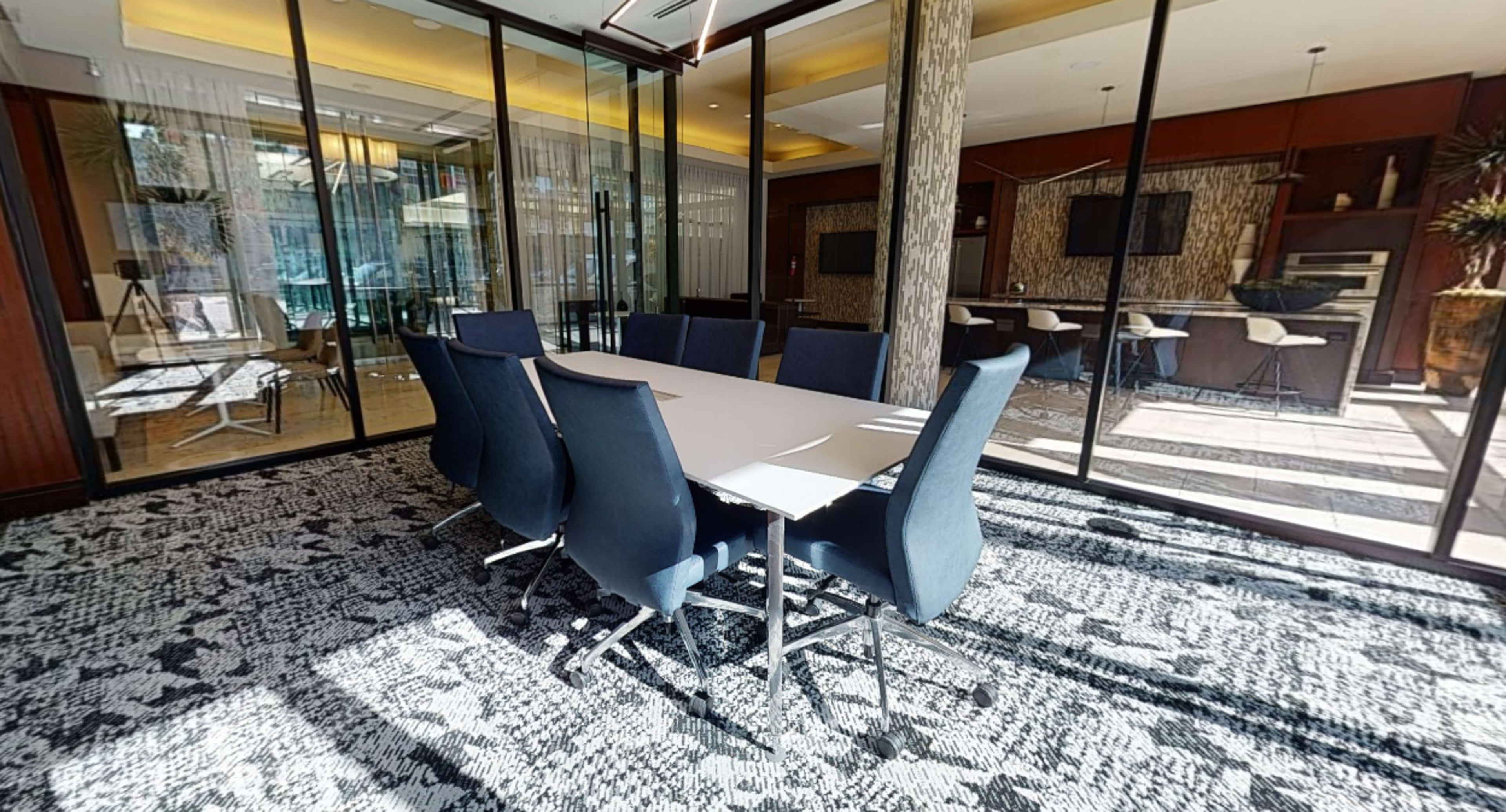 A conference room features a long table surrounded by eight blue chairs, with large glass windows allowing natural light to fill the space.