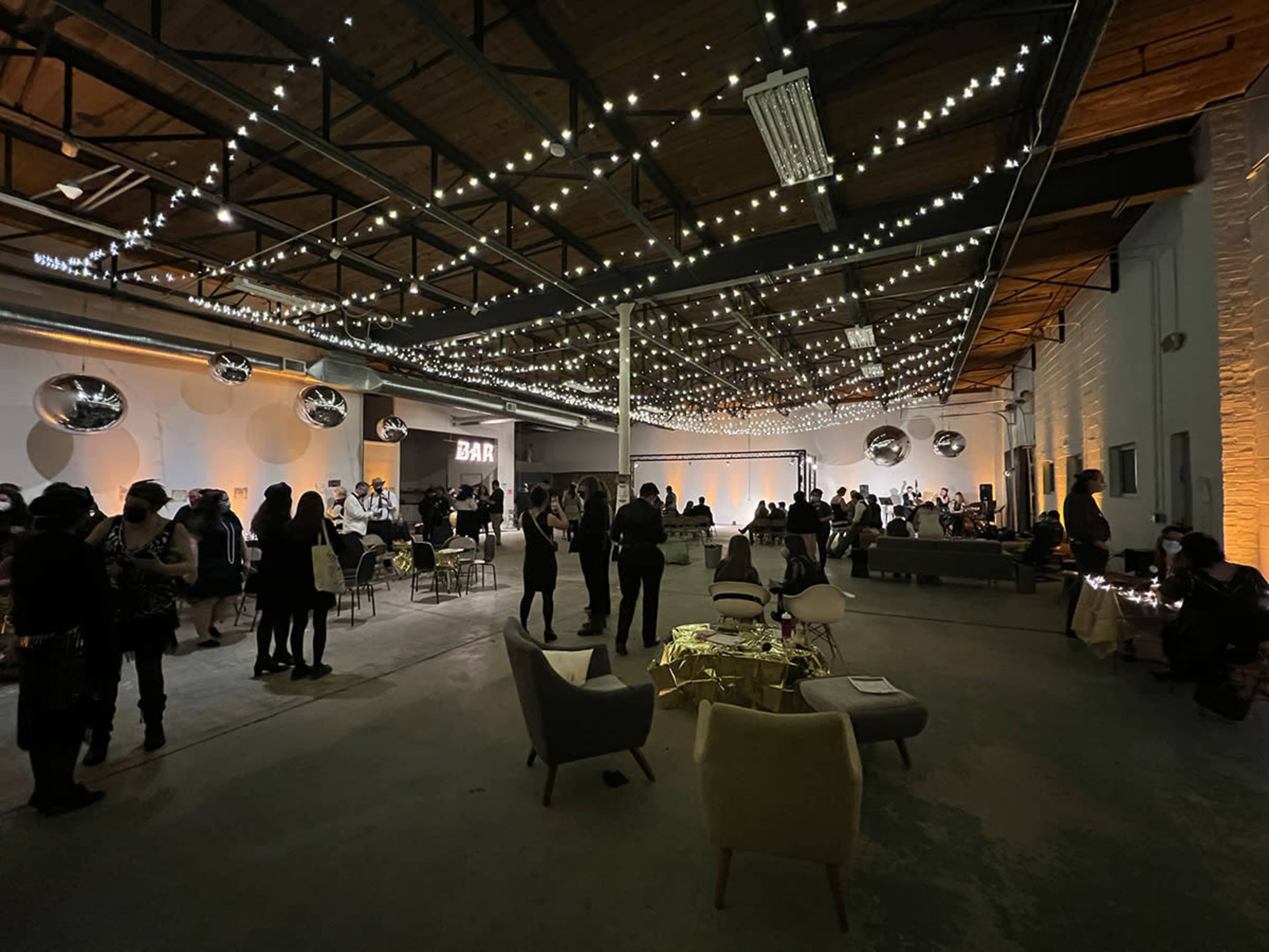A spacious event venue decorated with string lights, featuring seated guests mingling and a bar area visible in the background.