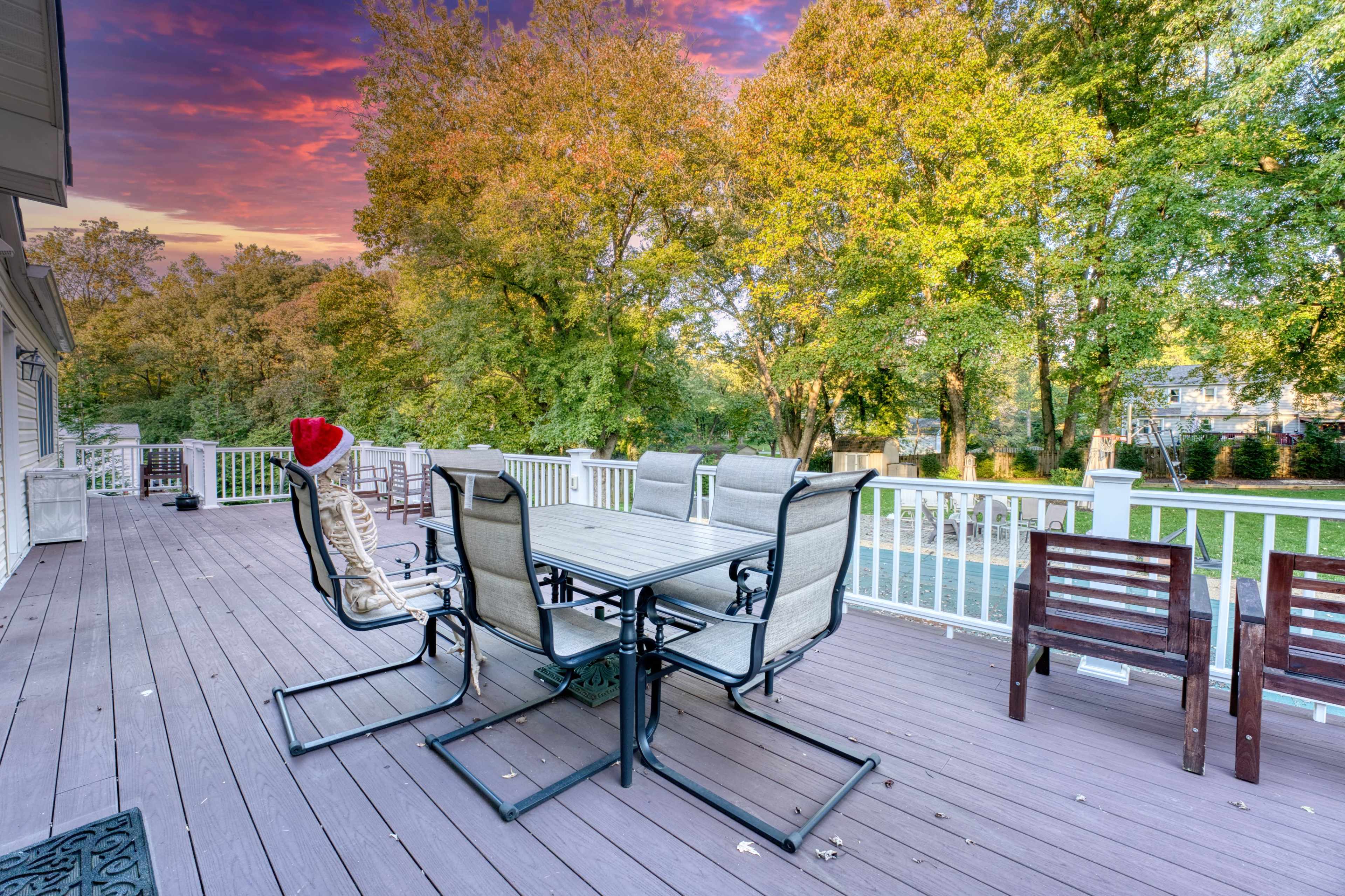 A patio with a dining table and chairs overlooks a wooded area with colorful autumn foliage under a sunset sky.