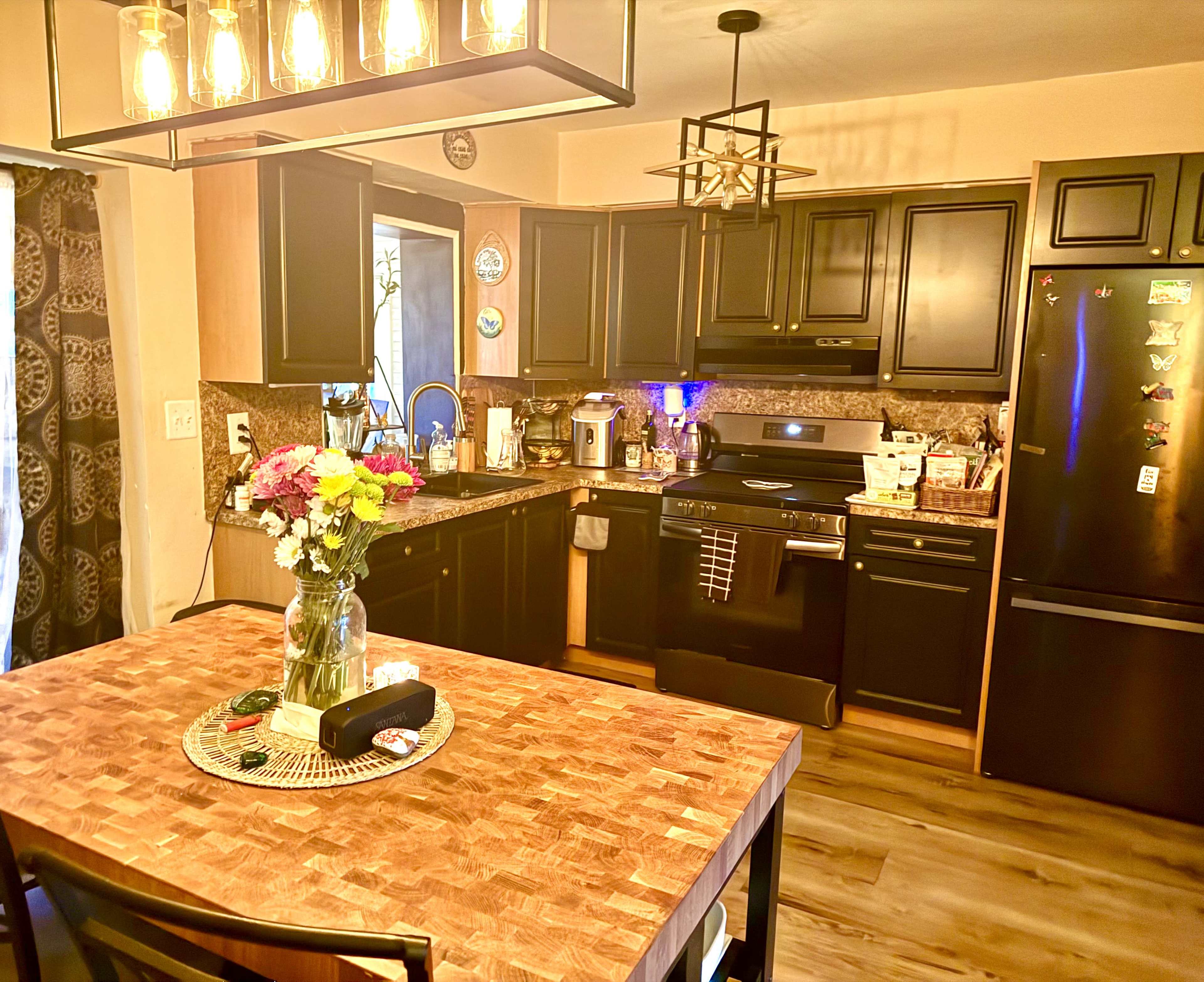 The image shows a modern kitchen with black cabinets, a wooden dining table, and various appliances arranged neatly on the countertops.