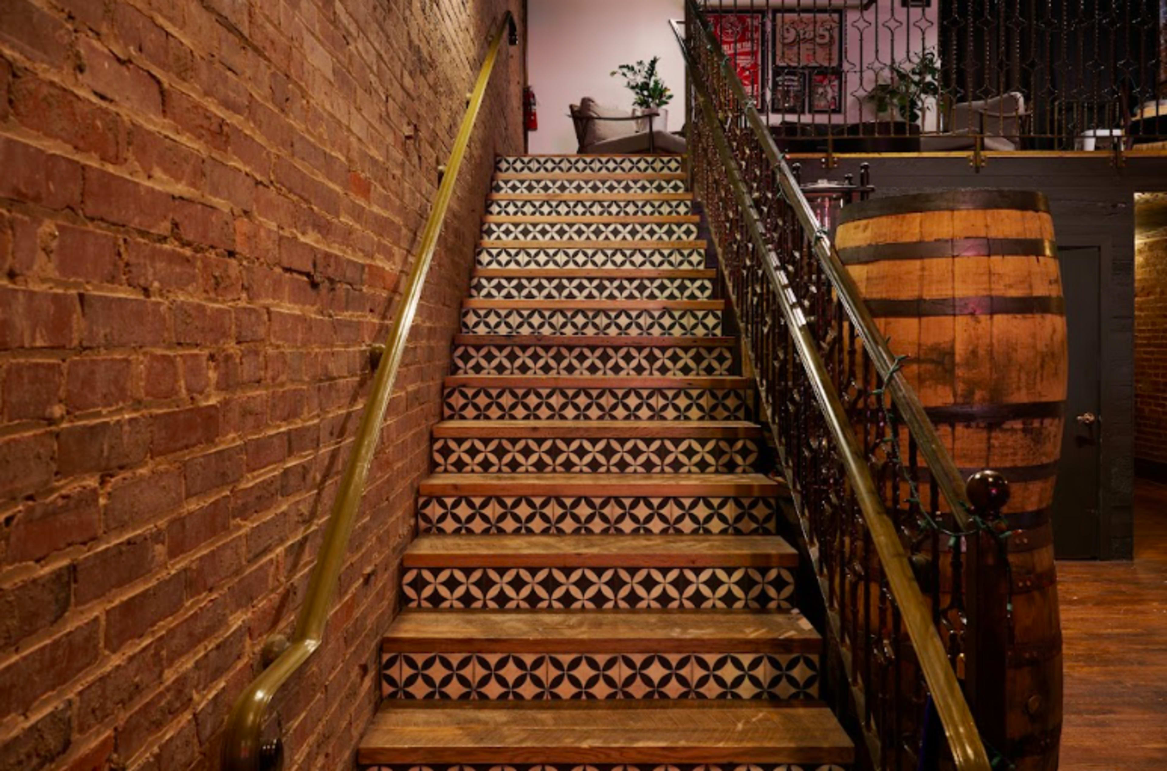 The image shows a staircase with patterned steps leading to an upper level, flanked by a brick wall and a wooden barrel on the side.
