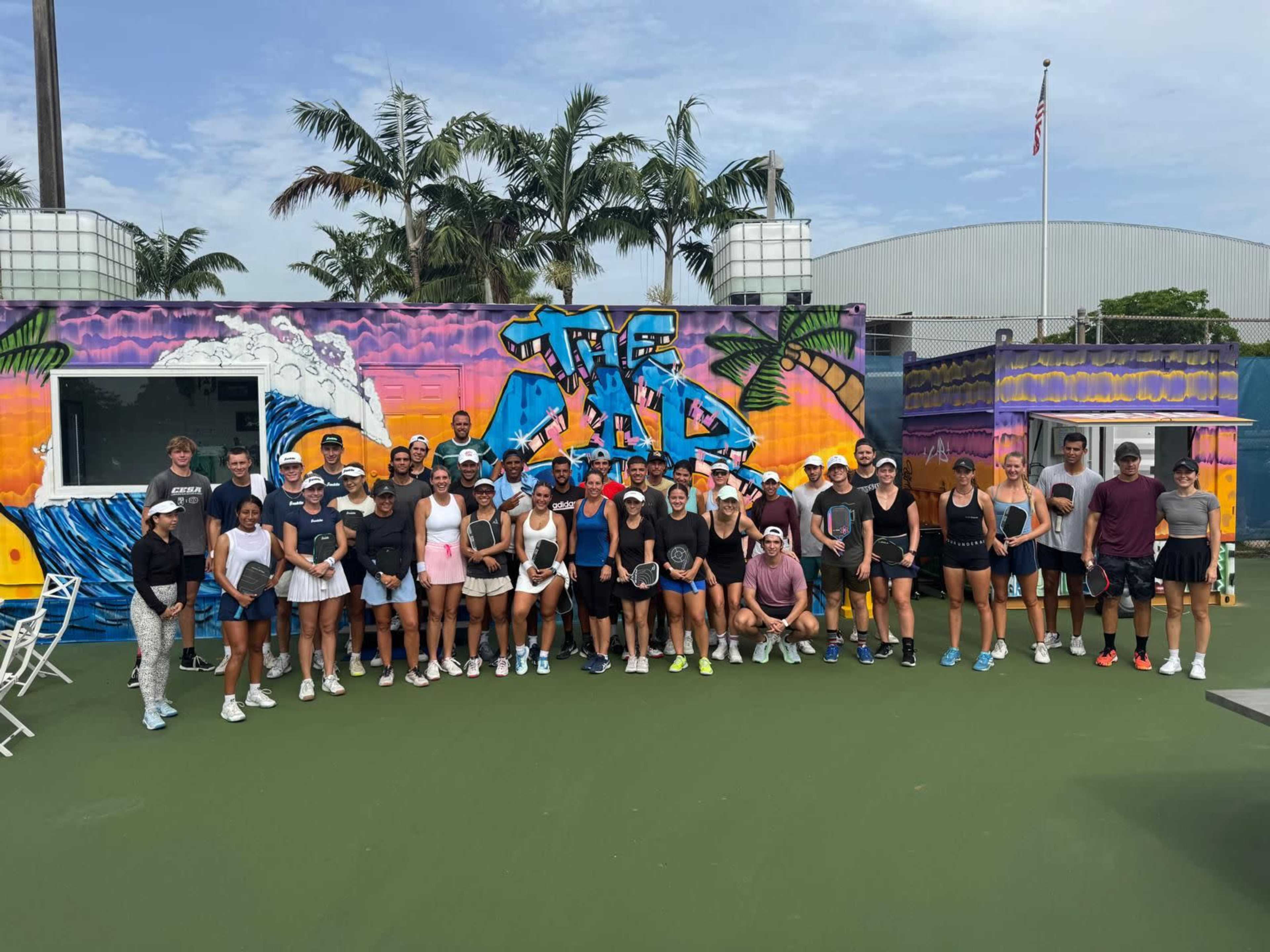 A large group of people is gathered in front of a colorful graffiti mural at a tennis facility, with tennis equipment and a bright, sunny sky.
