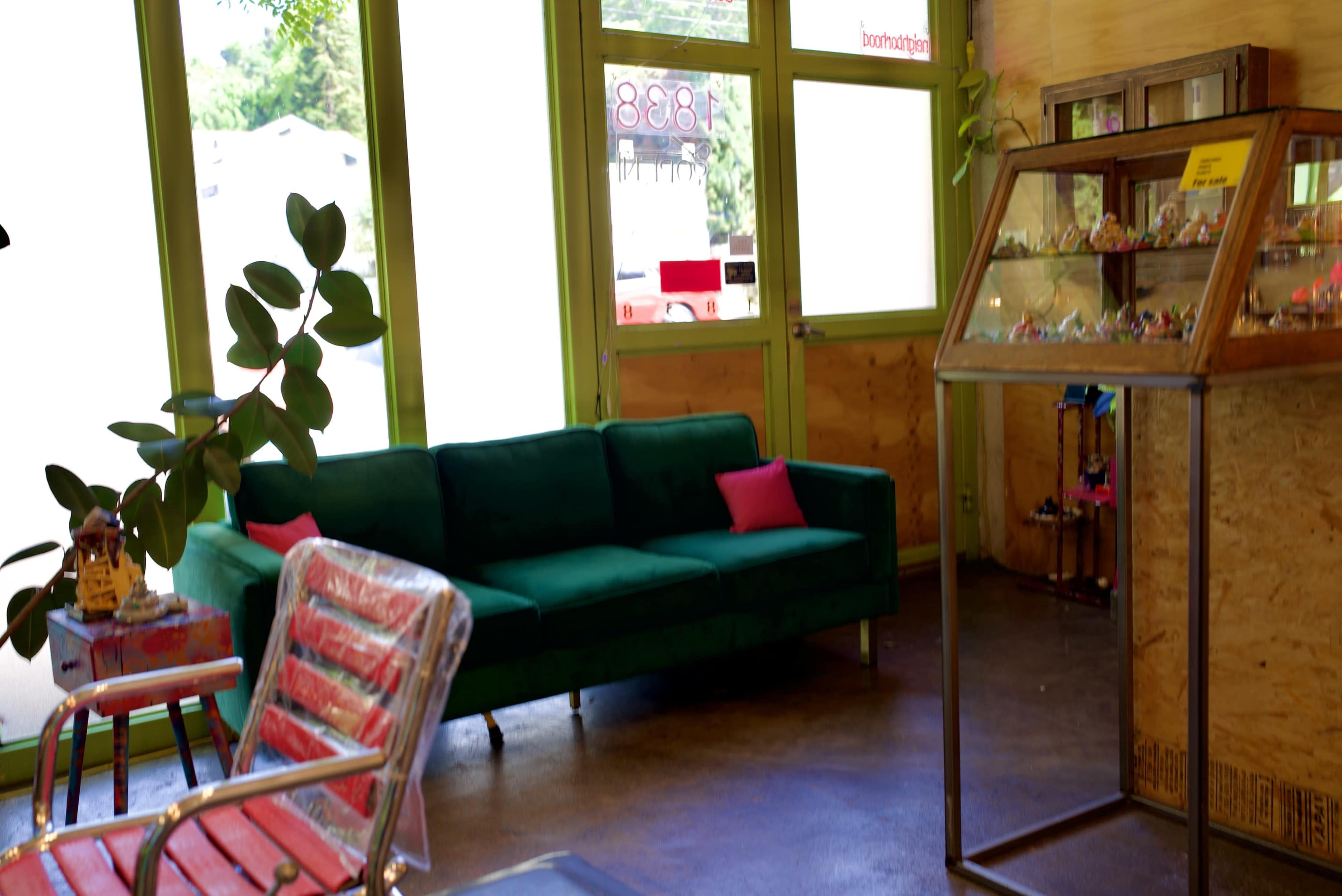 The image shows a modern interior with a green couch, a metal display case, and large windows allowing natural light to enter.