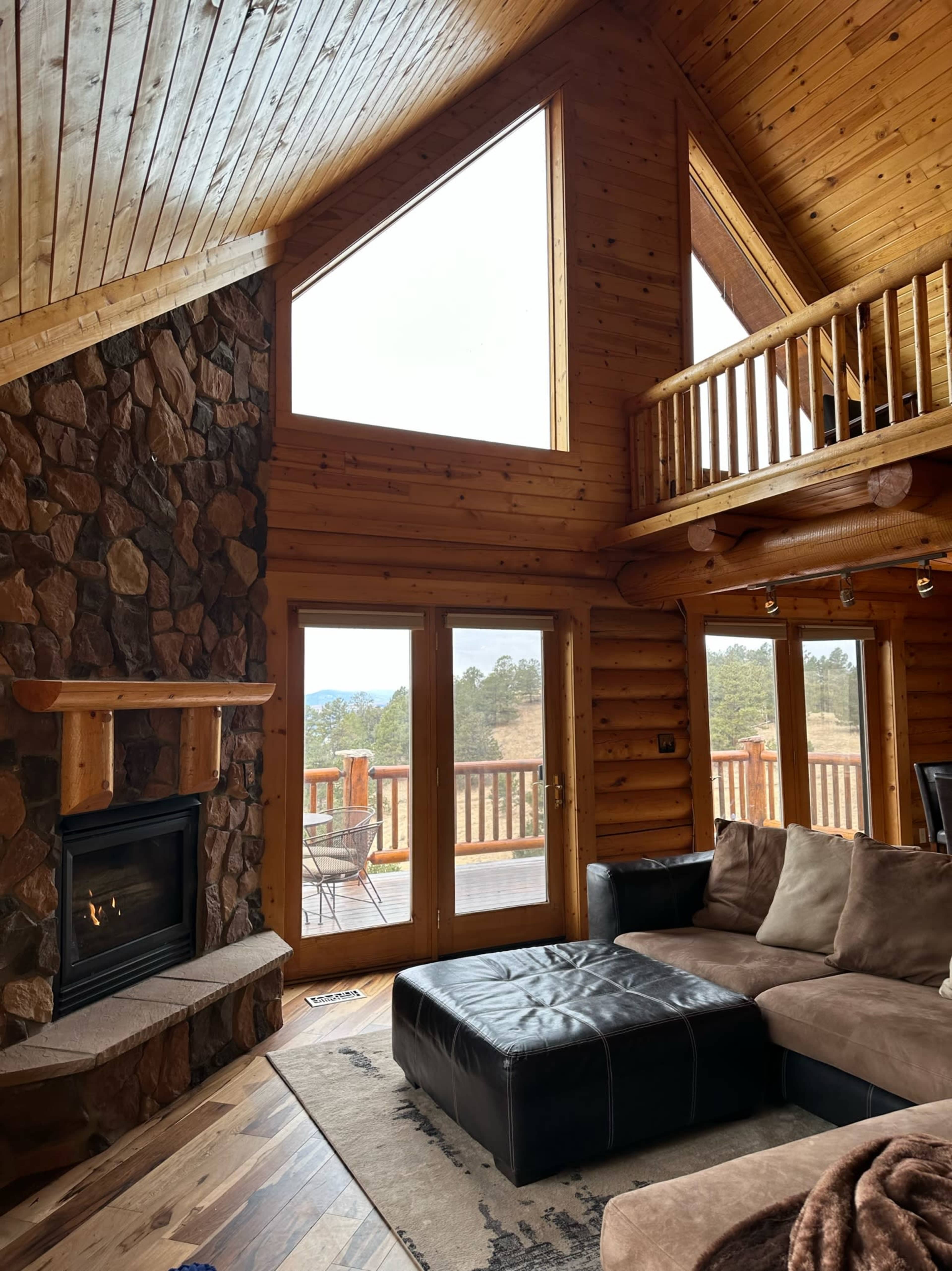A cozy wooden living room features a stone fireplace, large windows, and a sectional sofa, with access to a deck overlooking a scenic view.