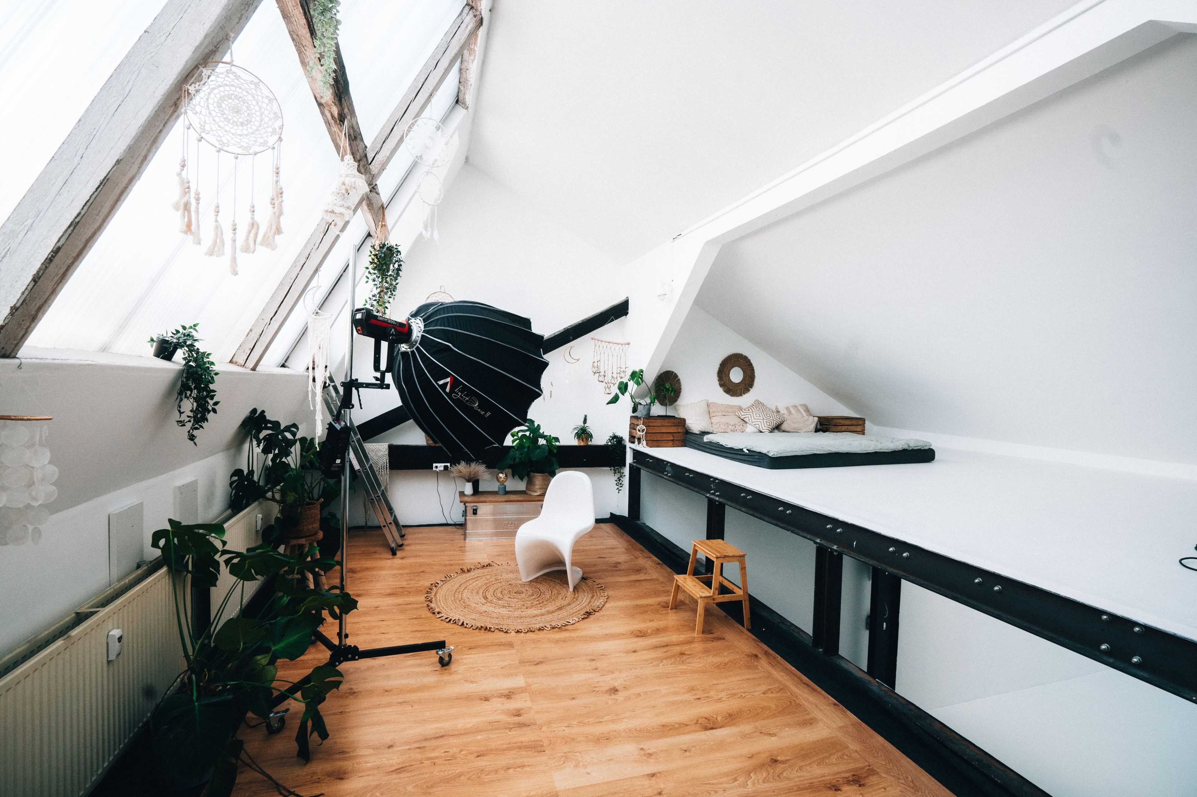 A bright, minimalist attic room with wooden flooring, a large softbox light, and a simple seating area surrounded by plants.
