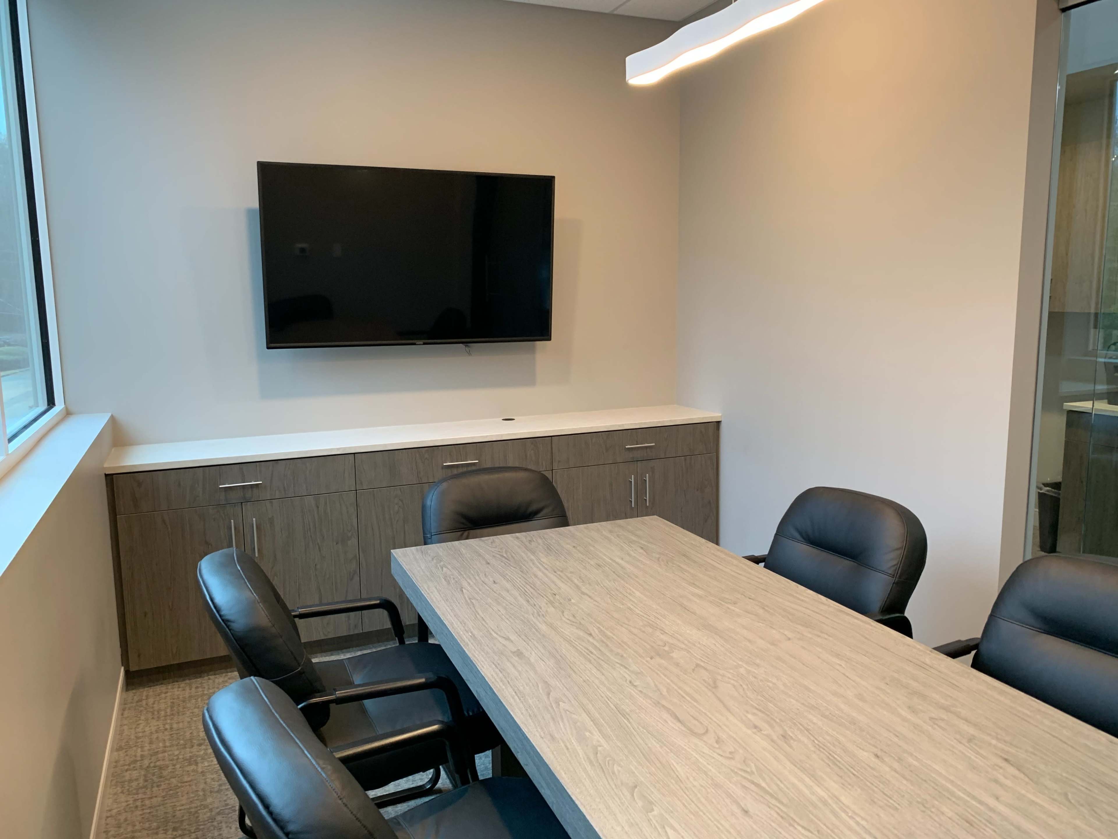 A conference room features a wooden table surrounded by black chairs, a mounted television on the wall, and a window providing natural light.