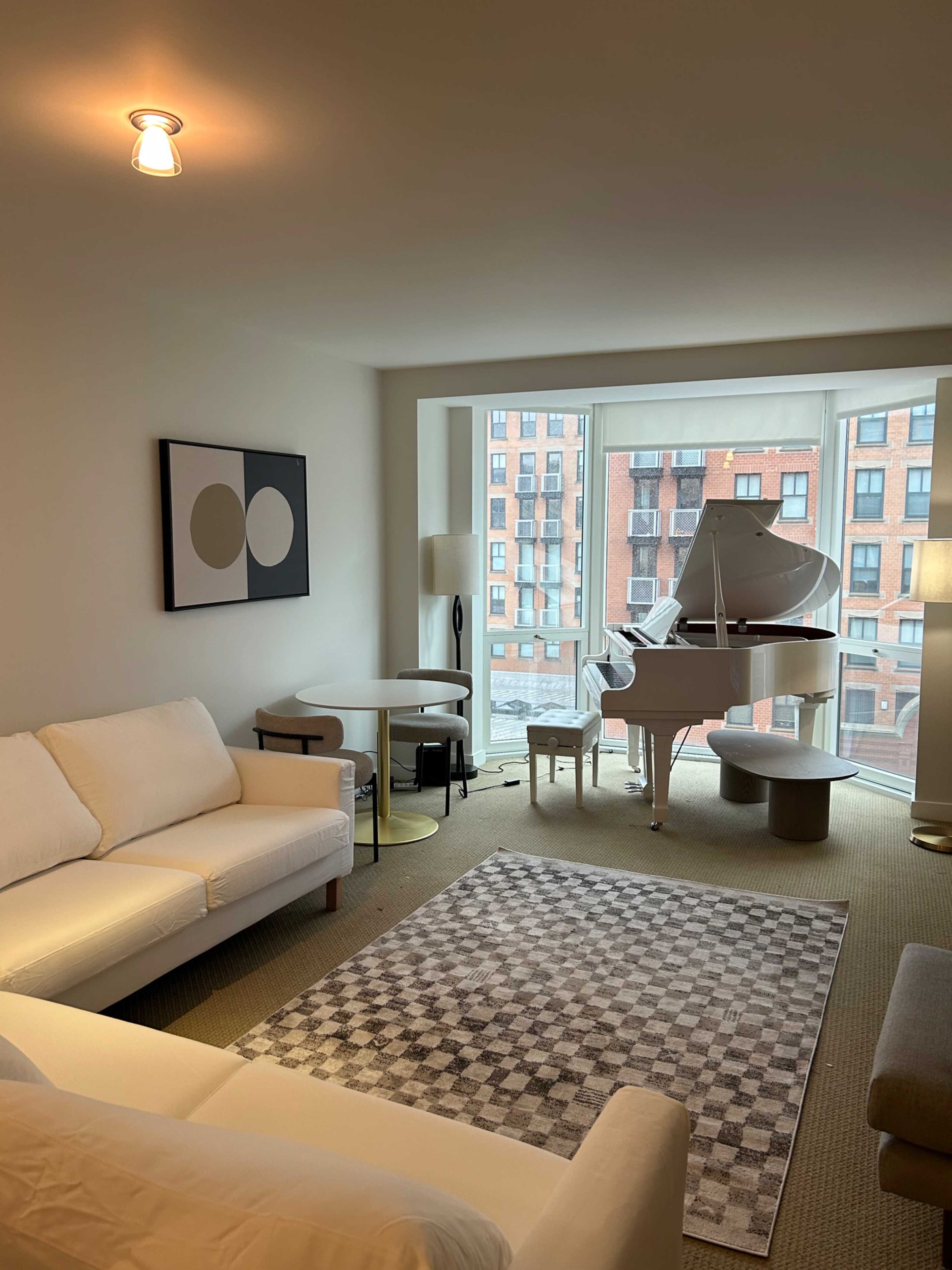 A modern living room features a white grand piano, a light sofa, a round table, and a gray patterned rug, with large windows showing a view of a brick building.