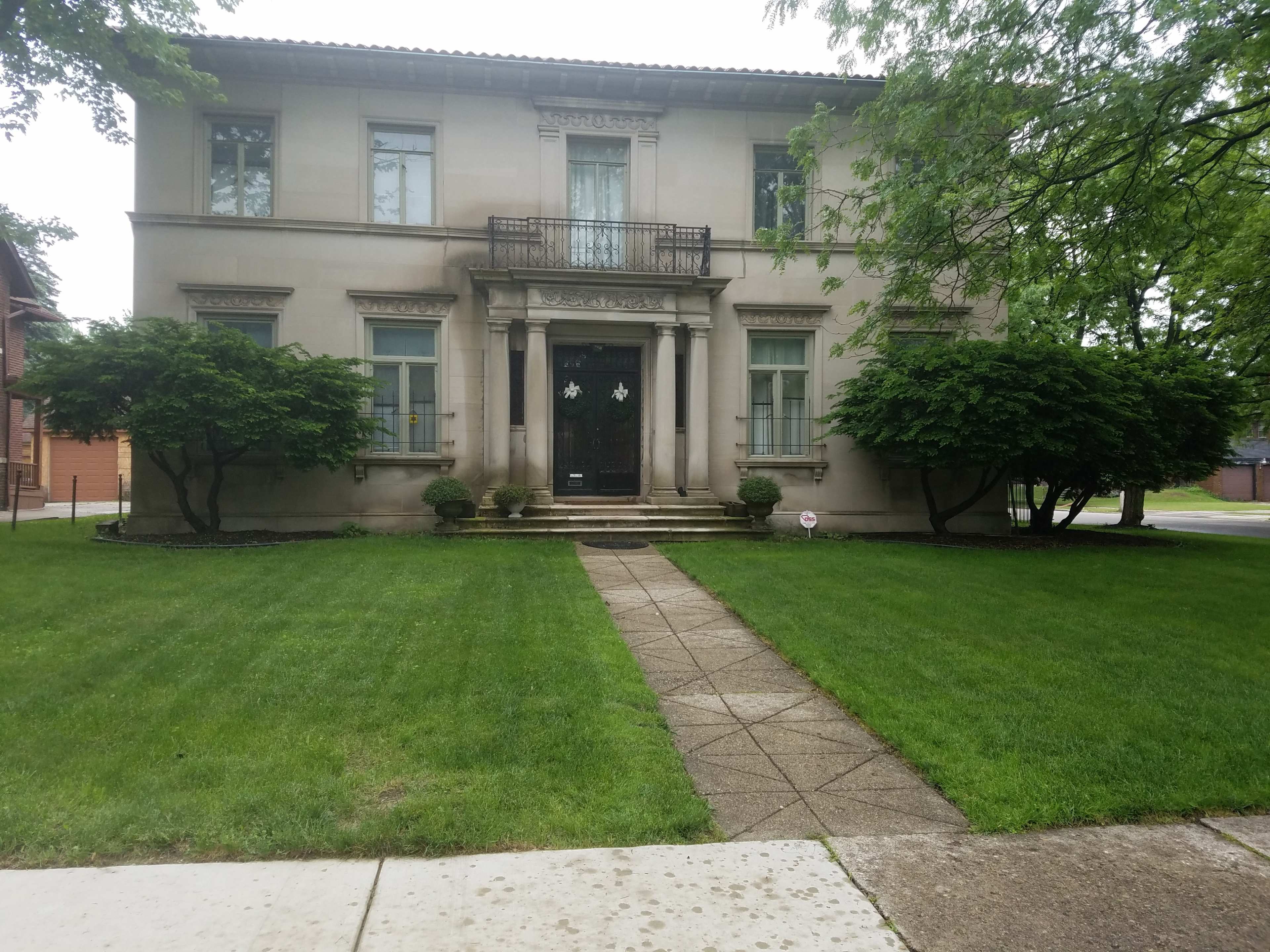 The image shows a large, two-story house with a symmetrical facade, a central staircase, and manicured shrubs flanking the entrance.