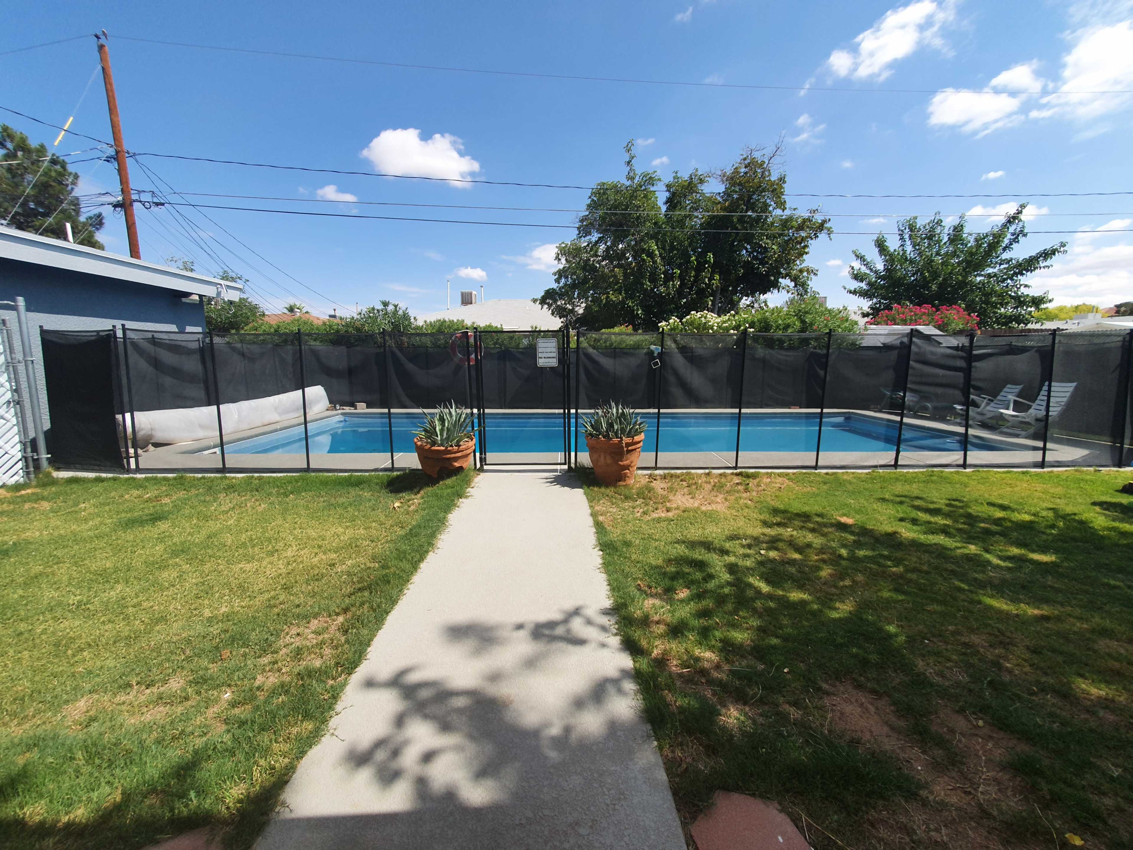 A fenced-in swimming pool surrounded by a green lawn and a concrete pathway leading towards it.