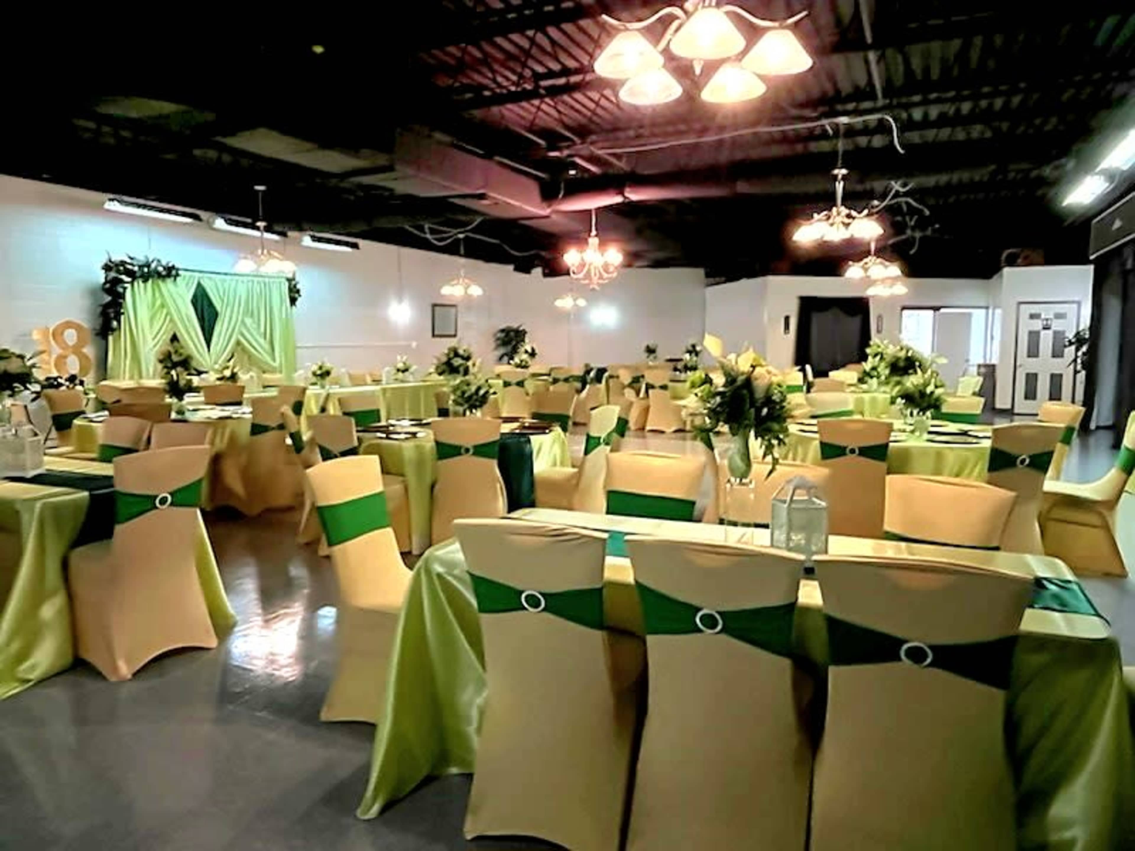The image shows a banquet hall set up with multiple tables covered in green and gold tablecloths, adorned with floral centerpieces and elegant lighting fixtures.