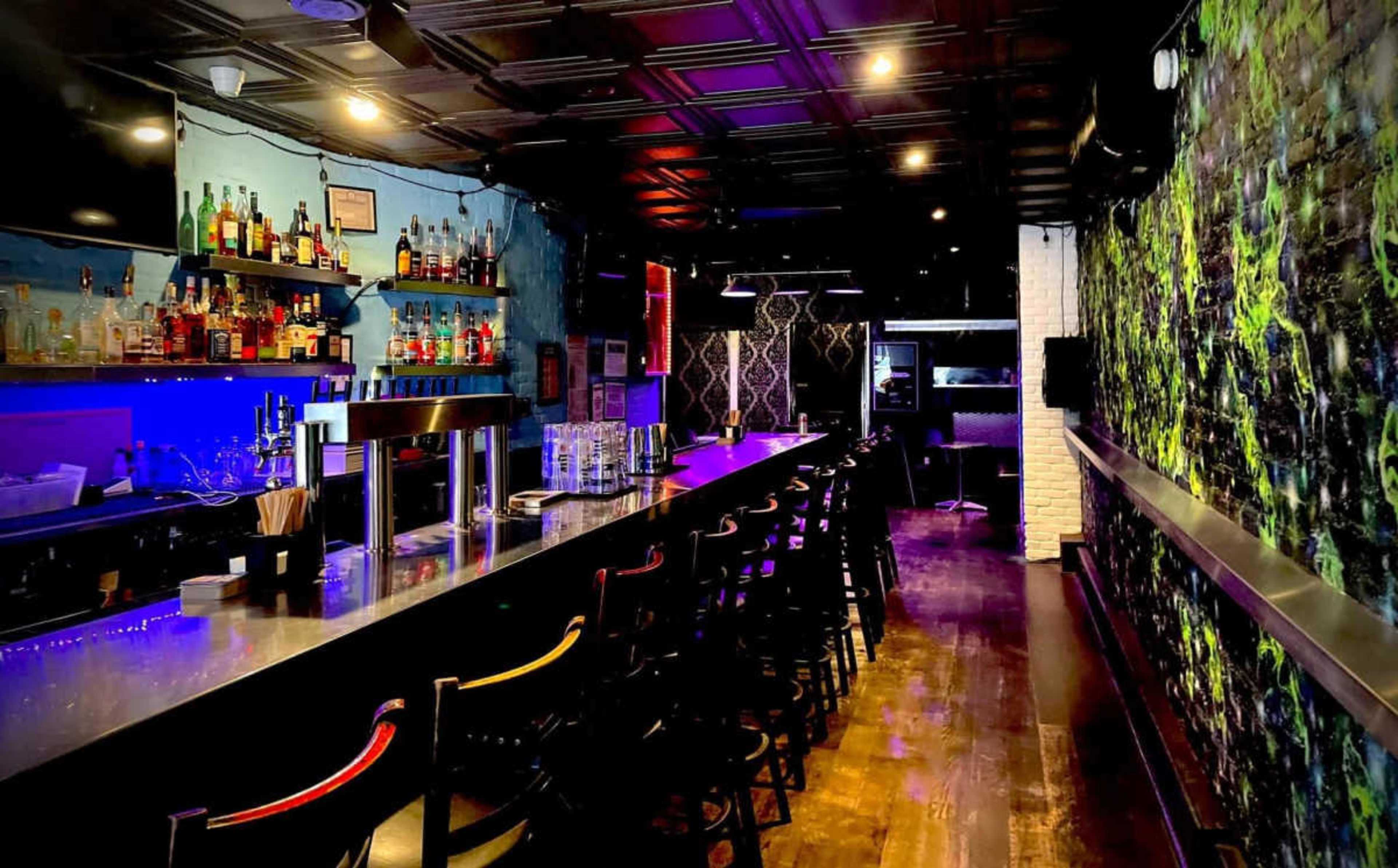 A dimly lit bar with a long countertop, shelves lined with various bottles, and a decorative wall featuring a vibrant design.