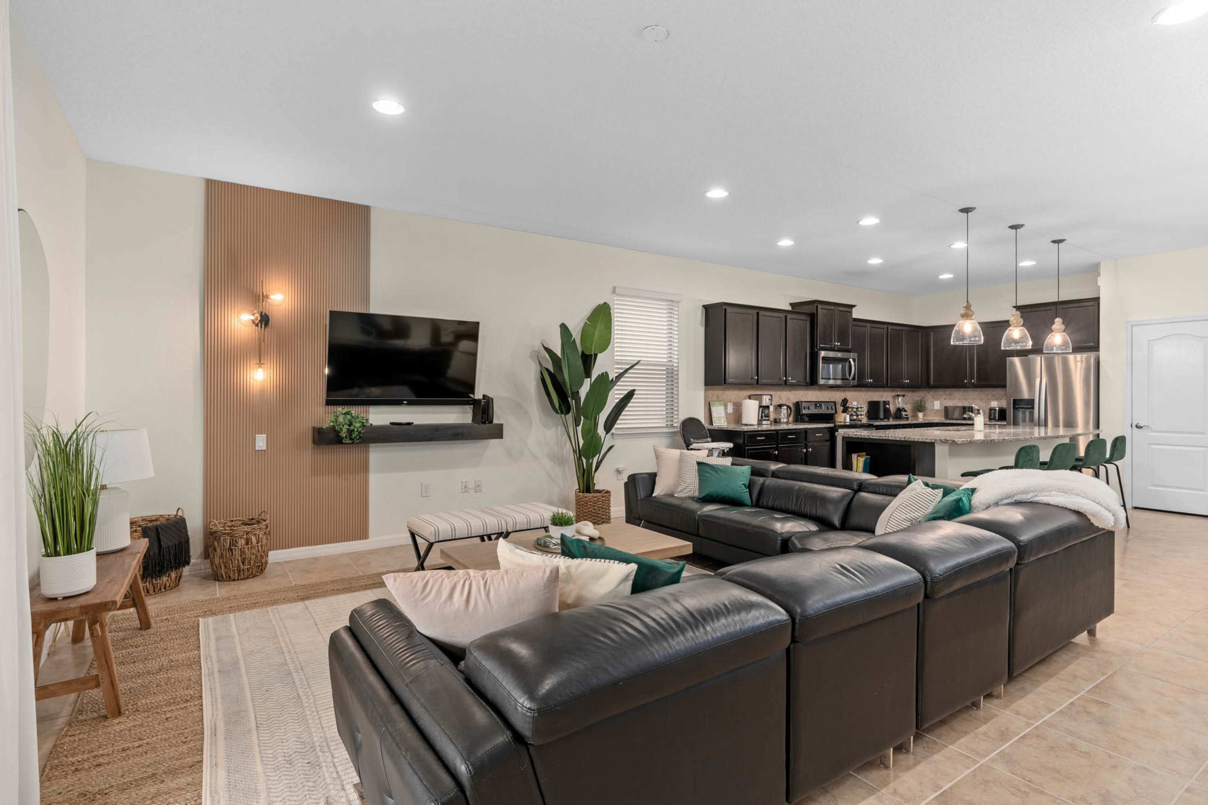 A modern living room with a dark leather sectional sofa, a wooden accent wall, and a kitchen area in the background featuring dark cabinetry and pendant lighting.