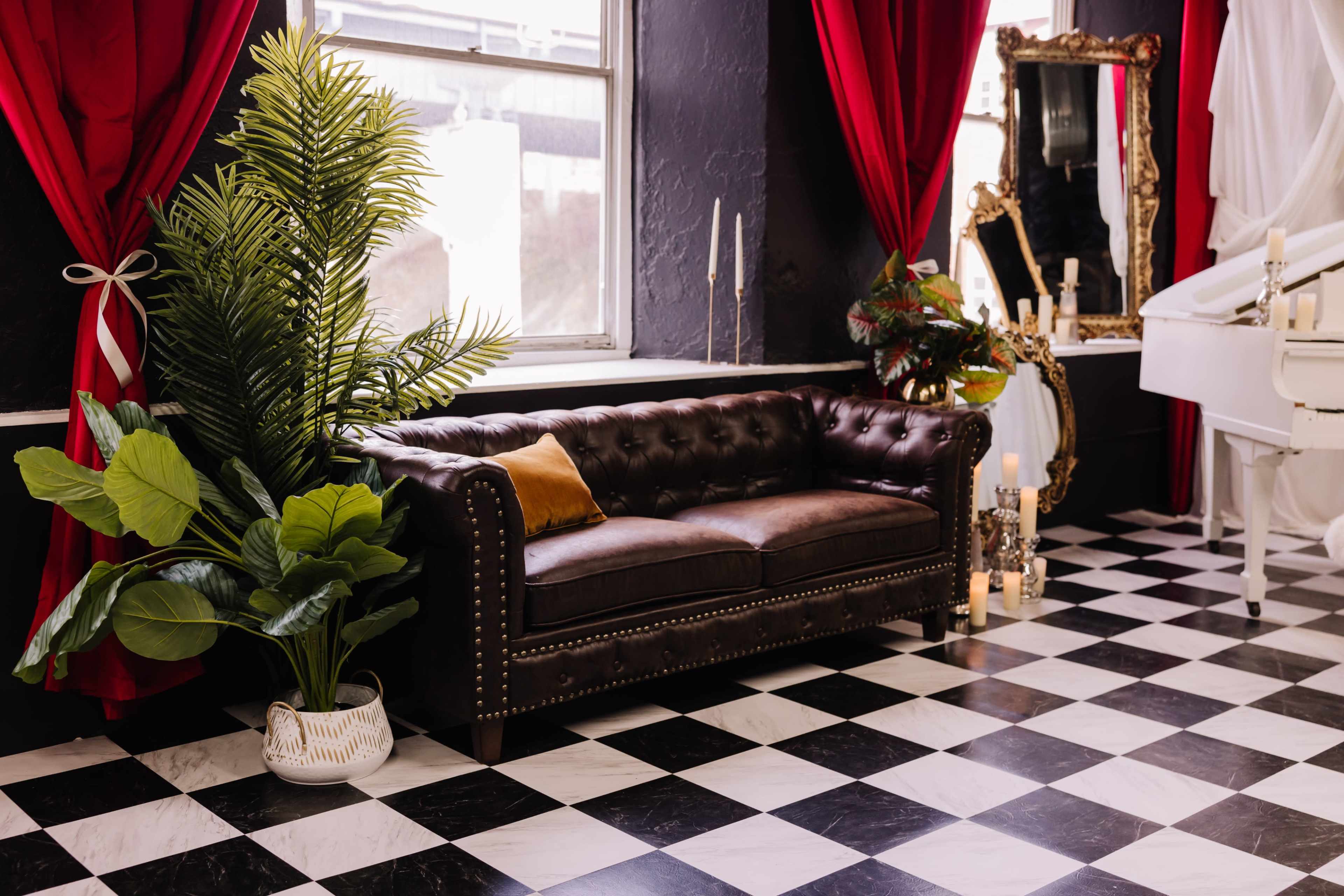 A brown leather chesterfield sofa with a decorative pillow is positioned beside large green plants in a room featuring black and white checkered flooring and red draped curtains.