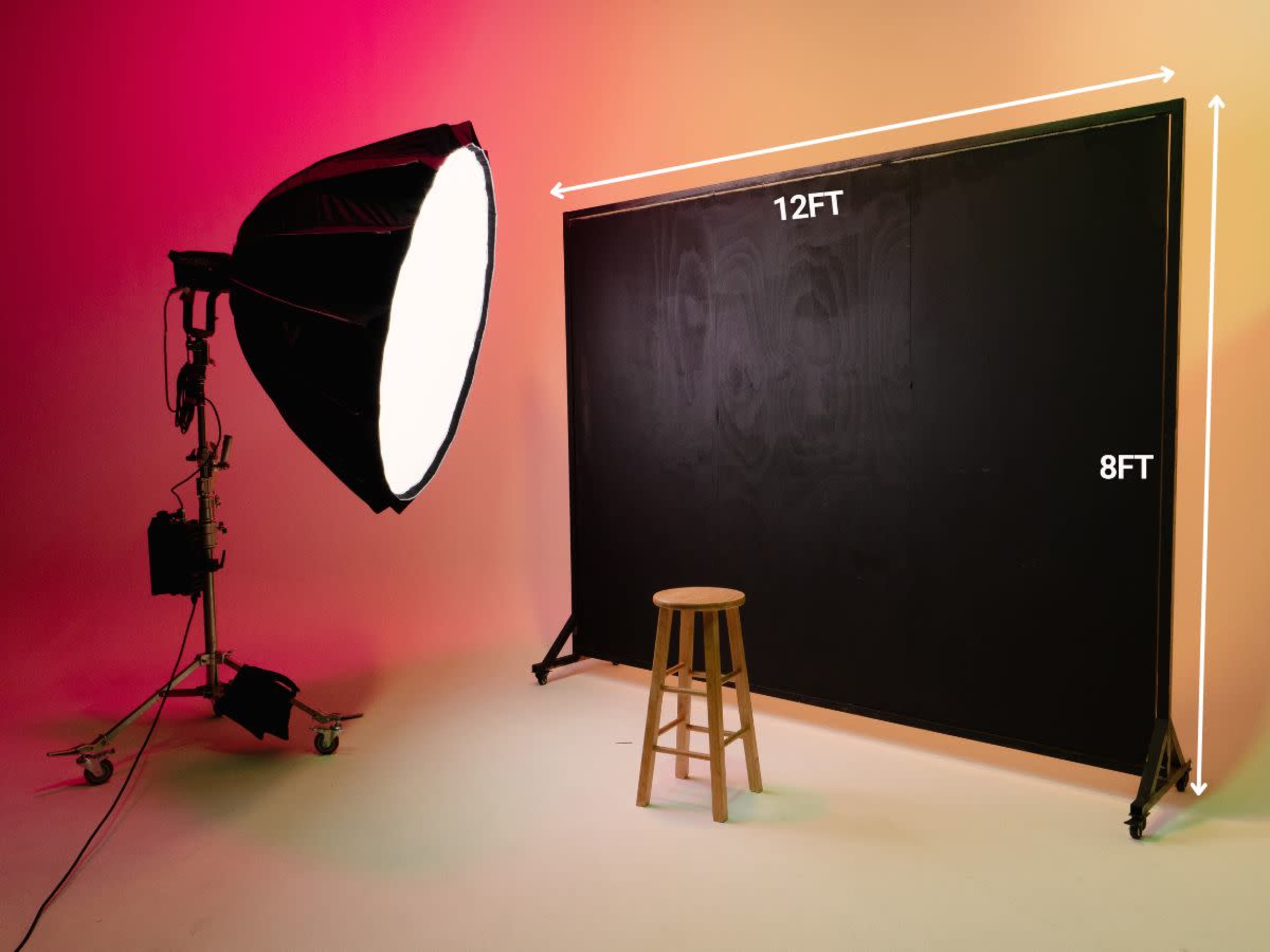 A photography setup featuring a large black backdrop measuring 12 feet wide and 8 feet tall, with a stool positioned in front and a softbox light on a stand to the side.