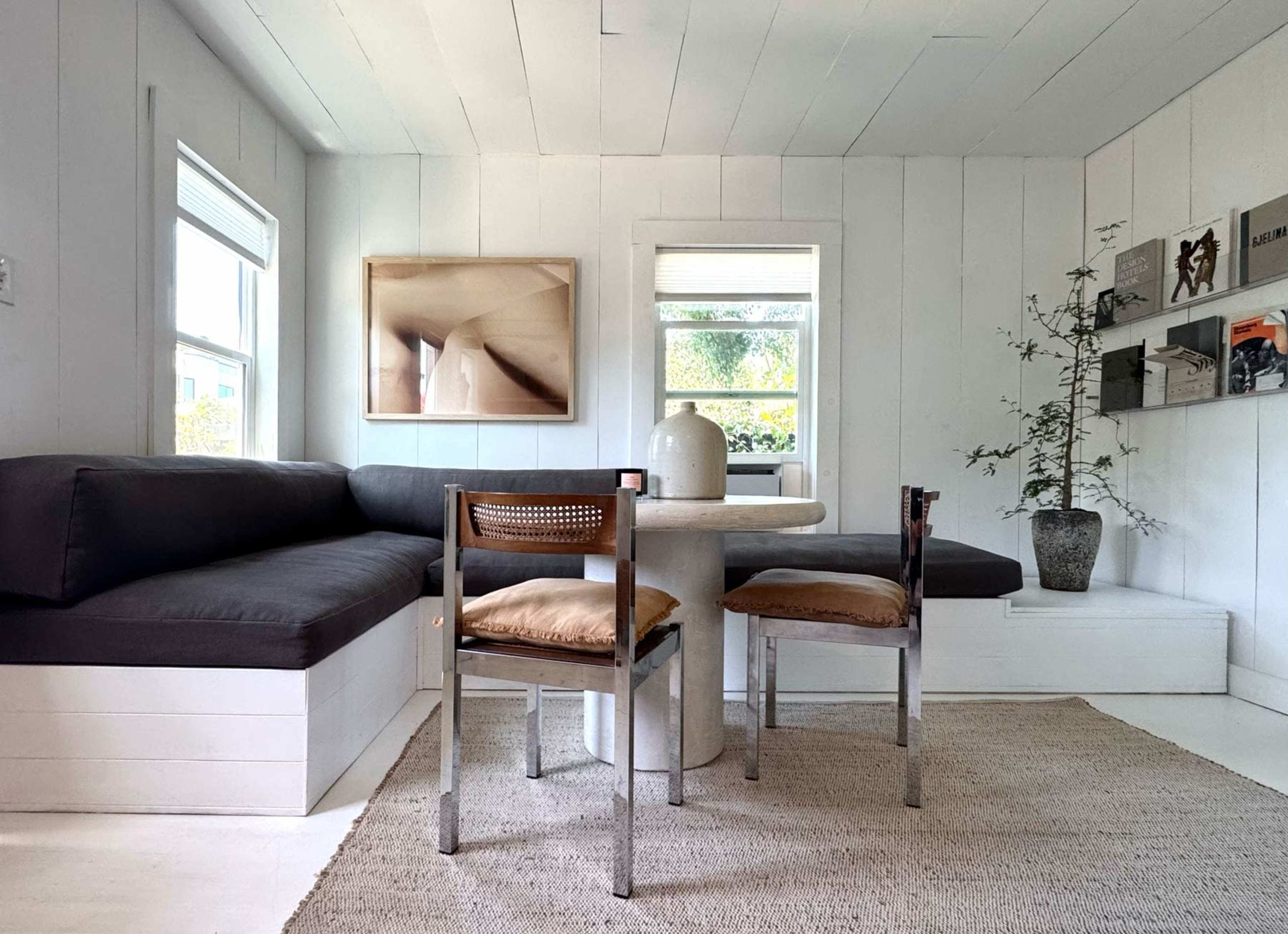 The image shows a minimalist, white-walled interior with a gray couch, a round table with a vase, and a small plant next to a shelf.