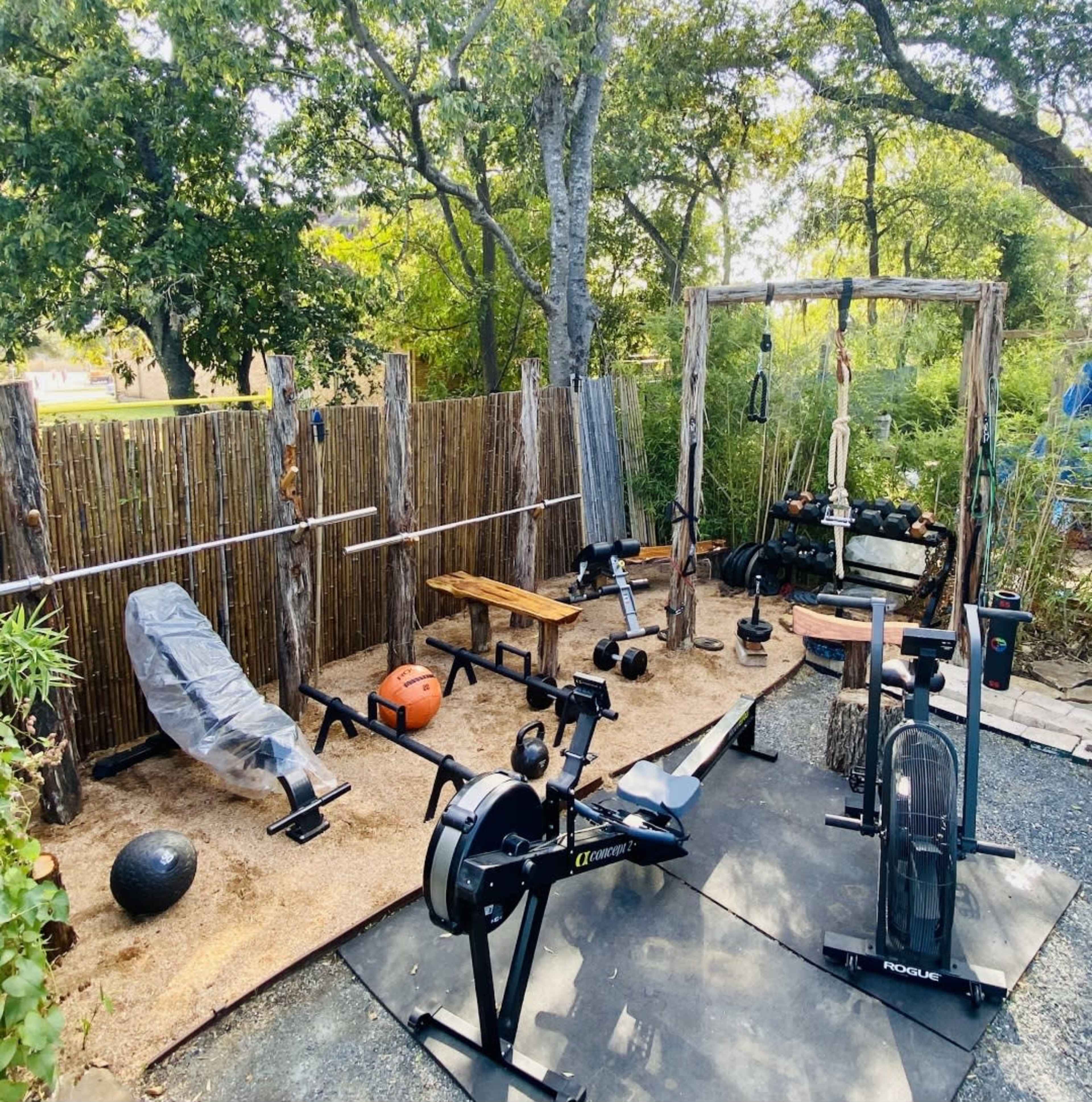 The image shows an outdoor workout area featuring various exercise equipment, including a rowing machine, free weights, and a bench, surrounded by natural wooden barriers.