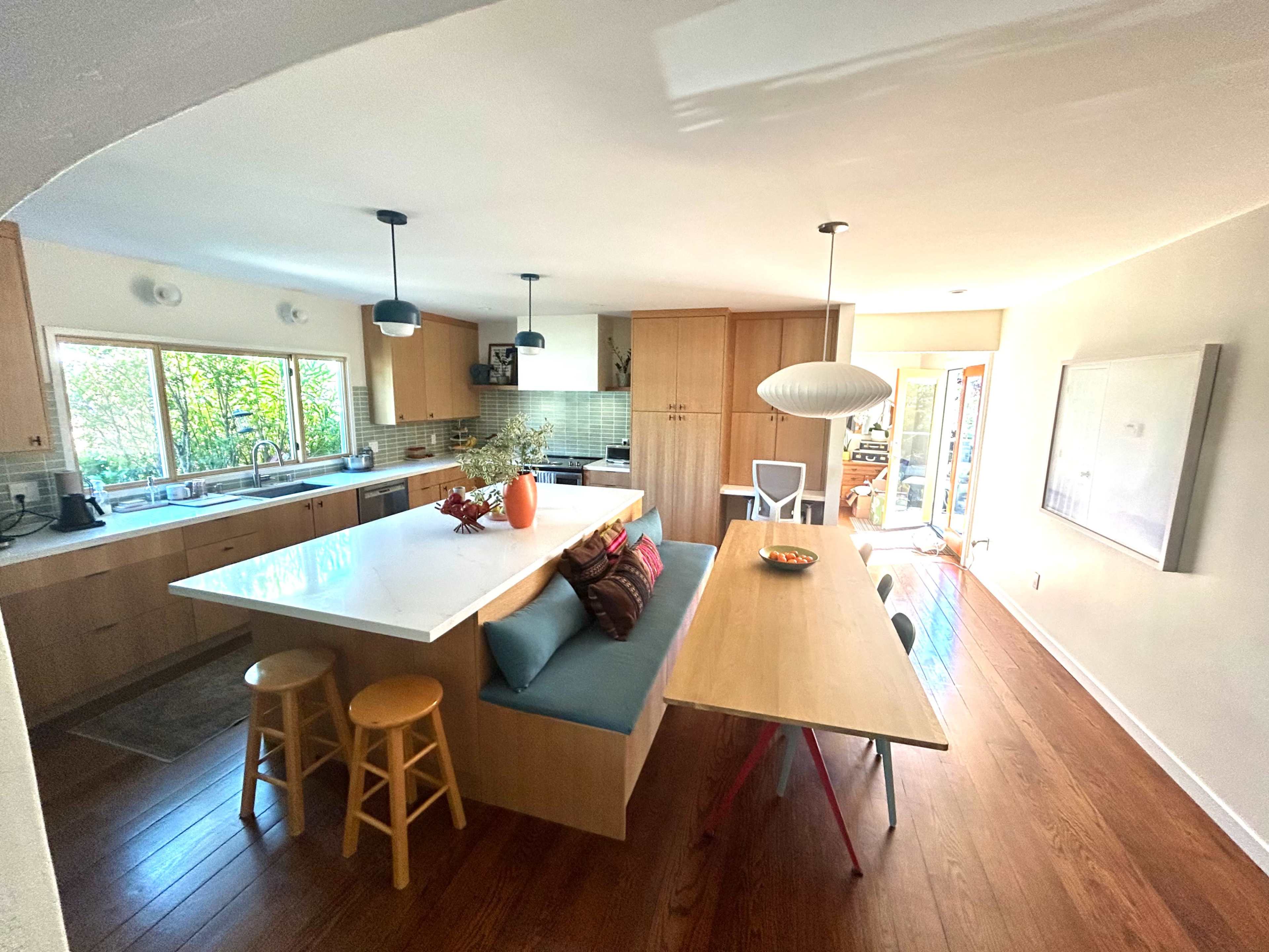 The image shows a modern kitchen and dining area featuring a large island with seating, a wooden dining table, and large windows allowing natural light to enter.
