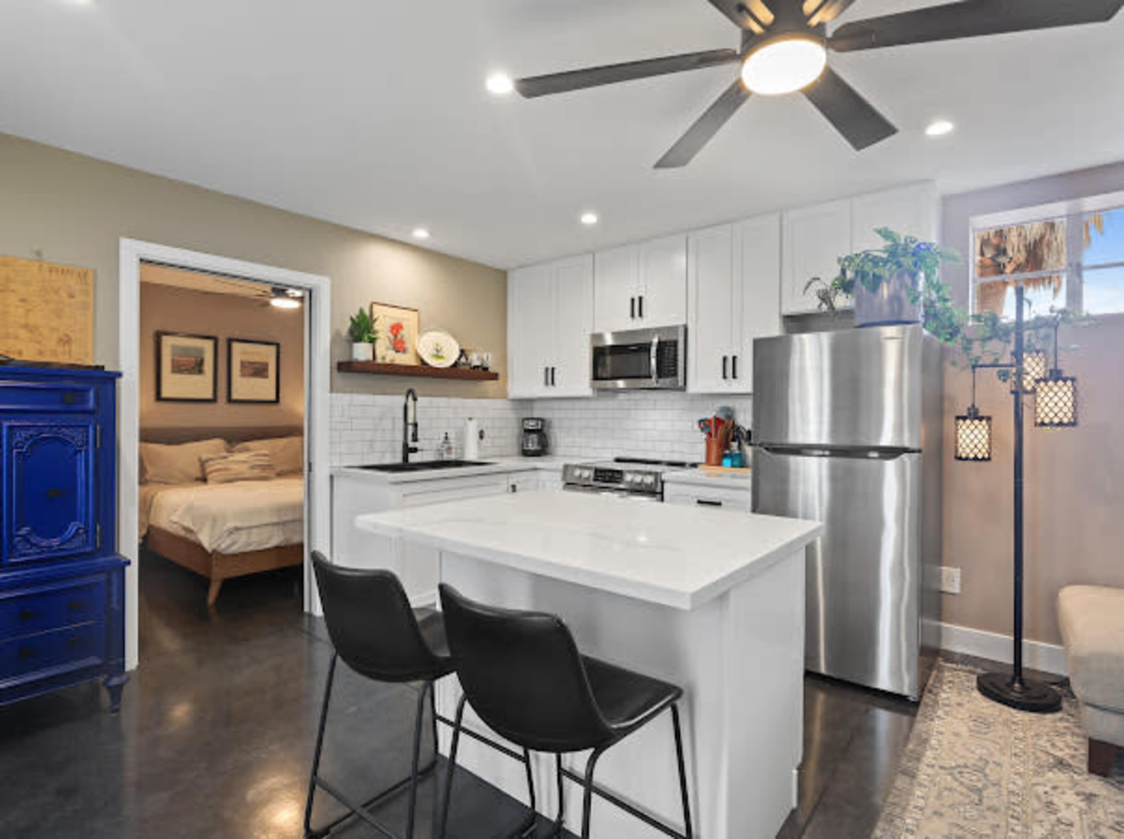The image shows a modern kitchen with white cabinetry, stainless steel appliances, a kitchen island with seating, and an adjoining living area visible through an open doorway.