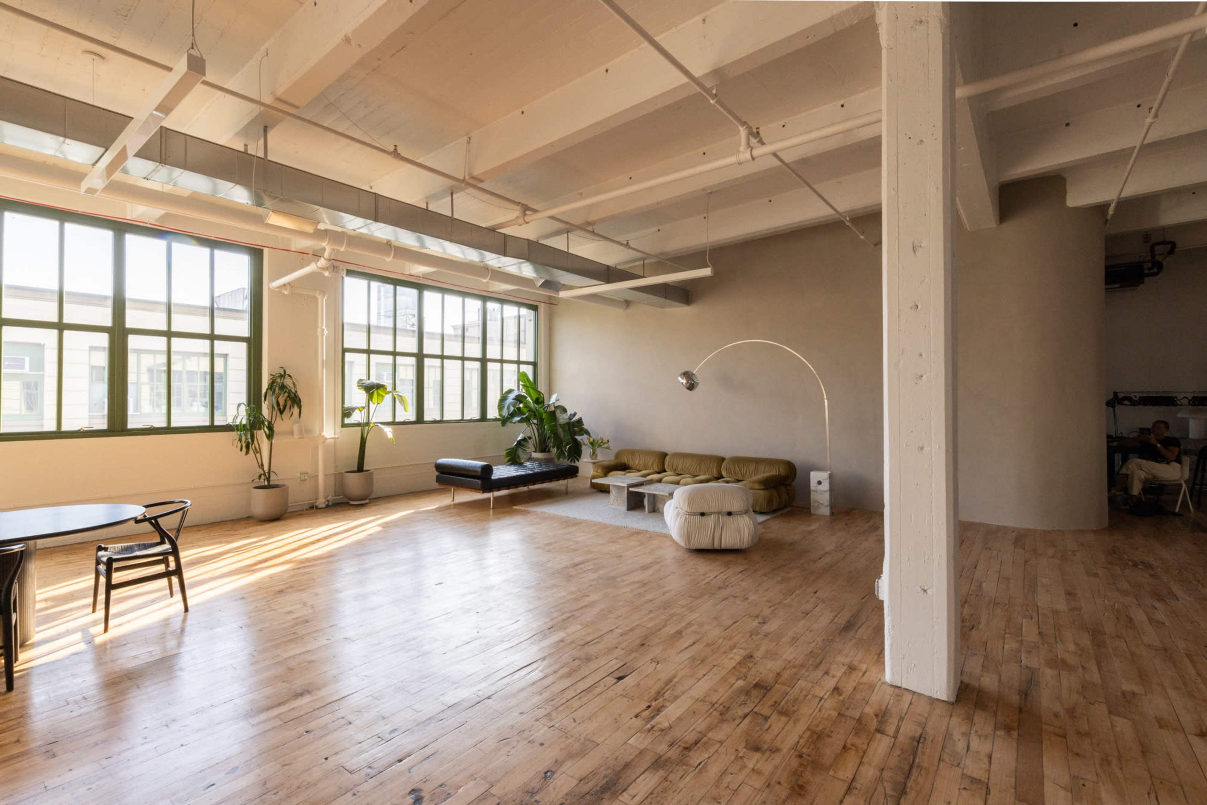 The image shows a spacious, well-lit room featuring wooden floors, large windows, and minimalist furniture arranged with plants in the corners.