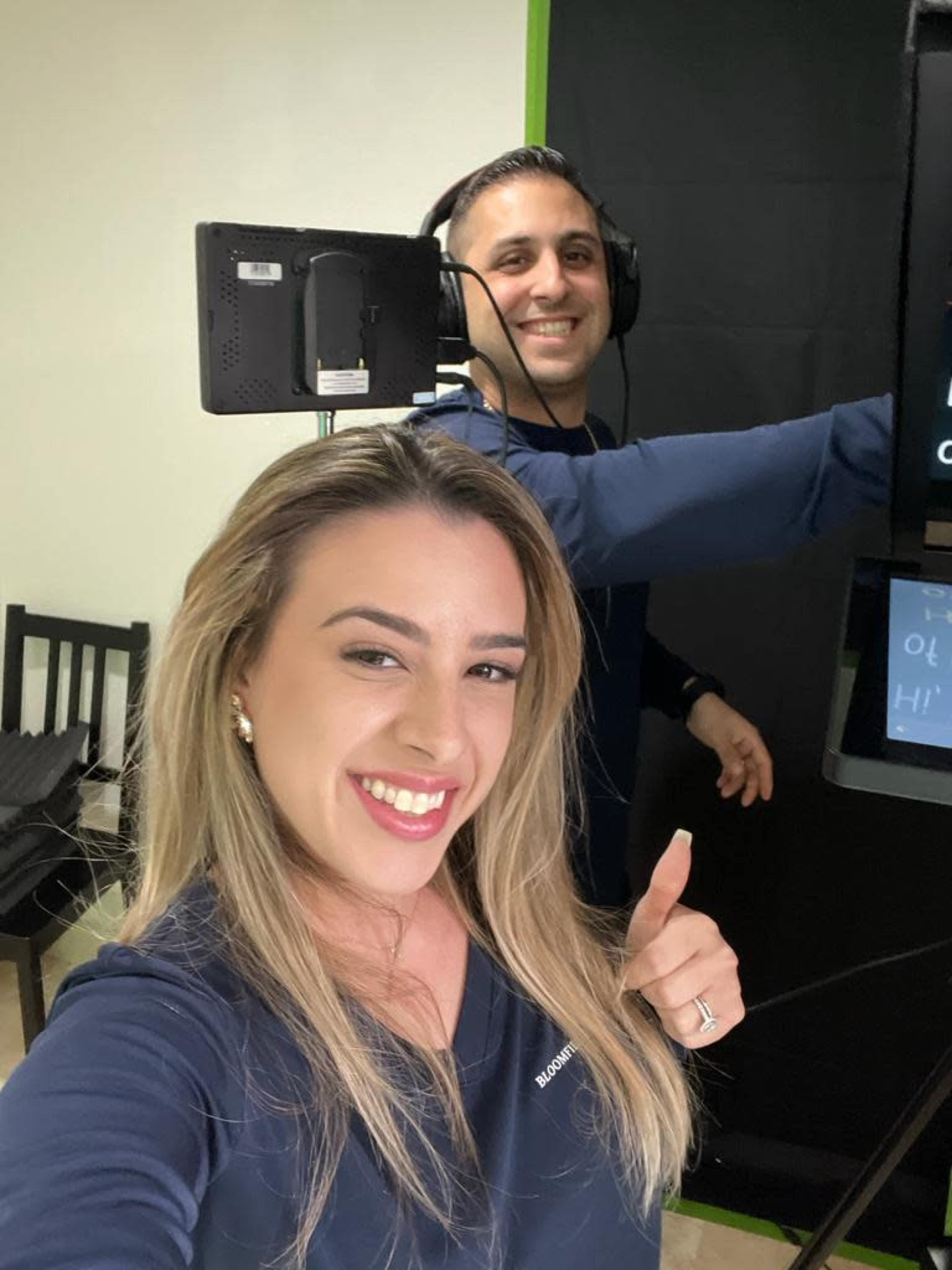 A woman smiles and gives a thumbs-up in the foreground while a man adjusts equipment in the background.