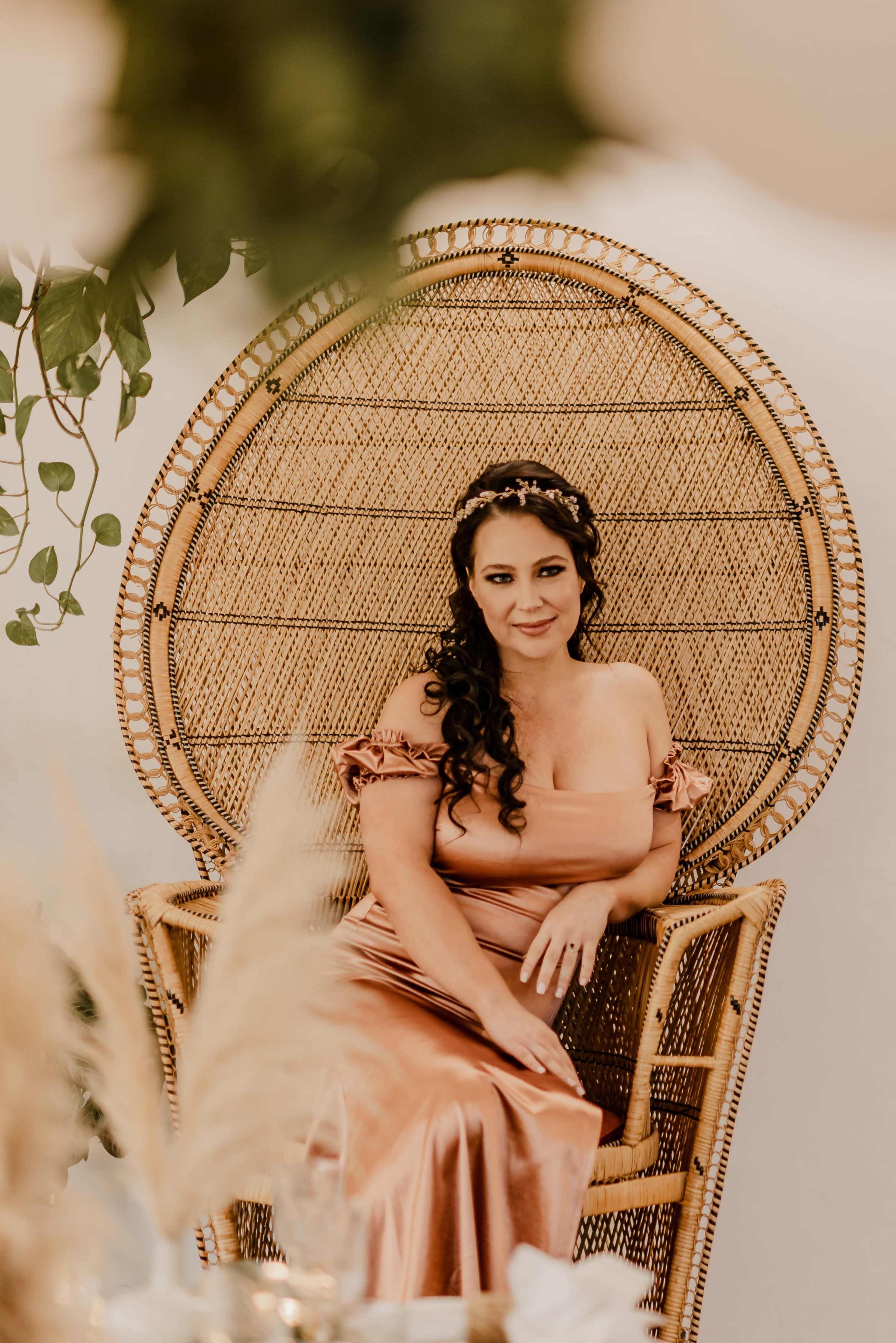 A woman in a bronze gown sits in a decorative woven chair against a light background.