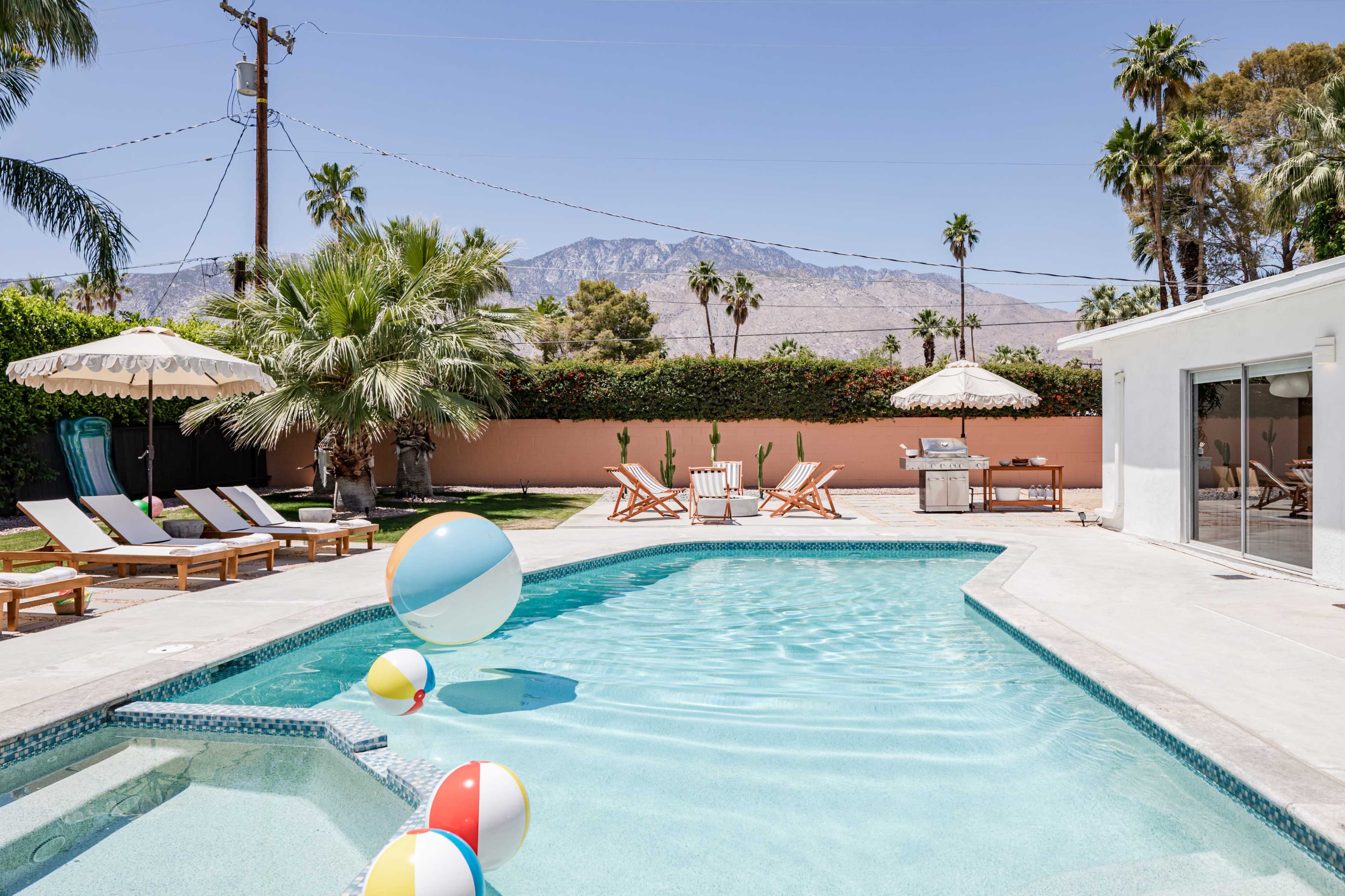 A sunny pool area features beach balls floating in the water, surrounded by lounge chairs, palm trees, and a backdrop of mountains.