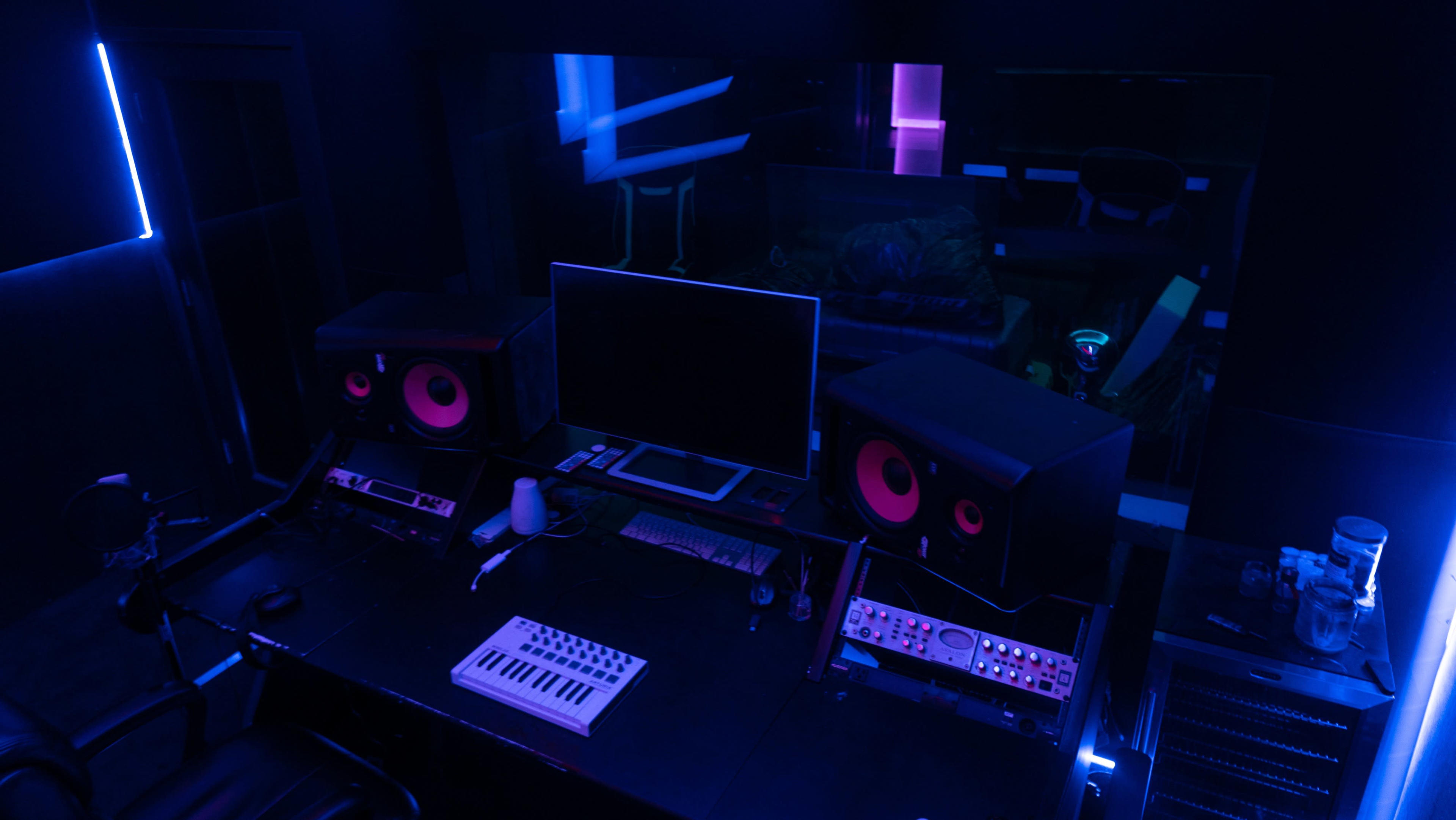 A dimly lit music studio featuring speakers, a computer monitor, and various music production equipment arranged on a black desk.