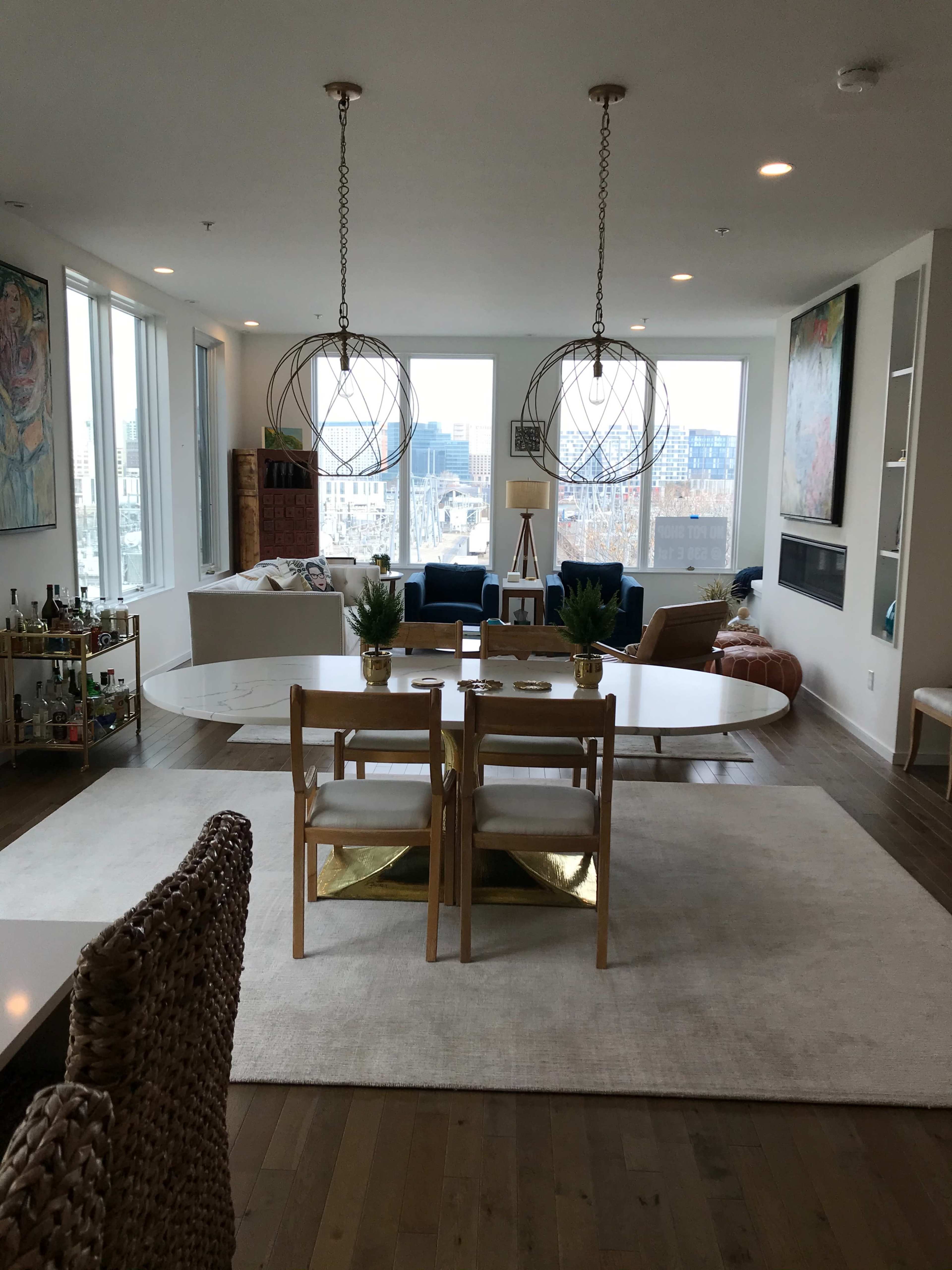 A modern living room features a large table surrounded by chairs, with large windows offering a view of the city and two spherical pendant lights hanging above.