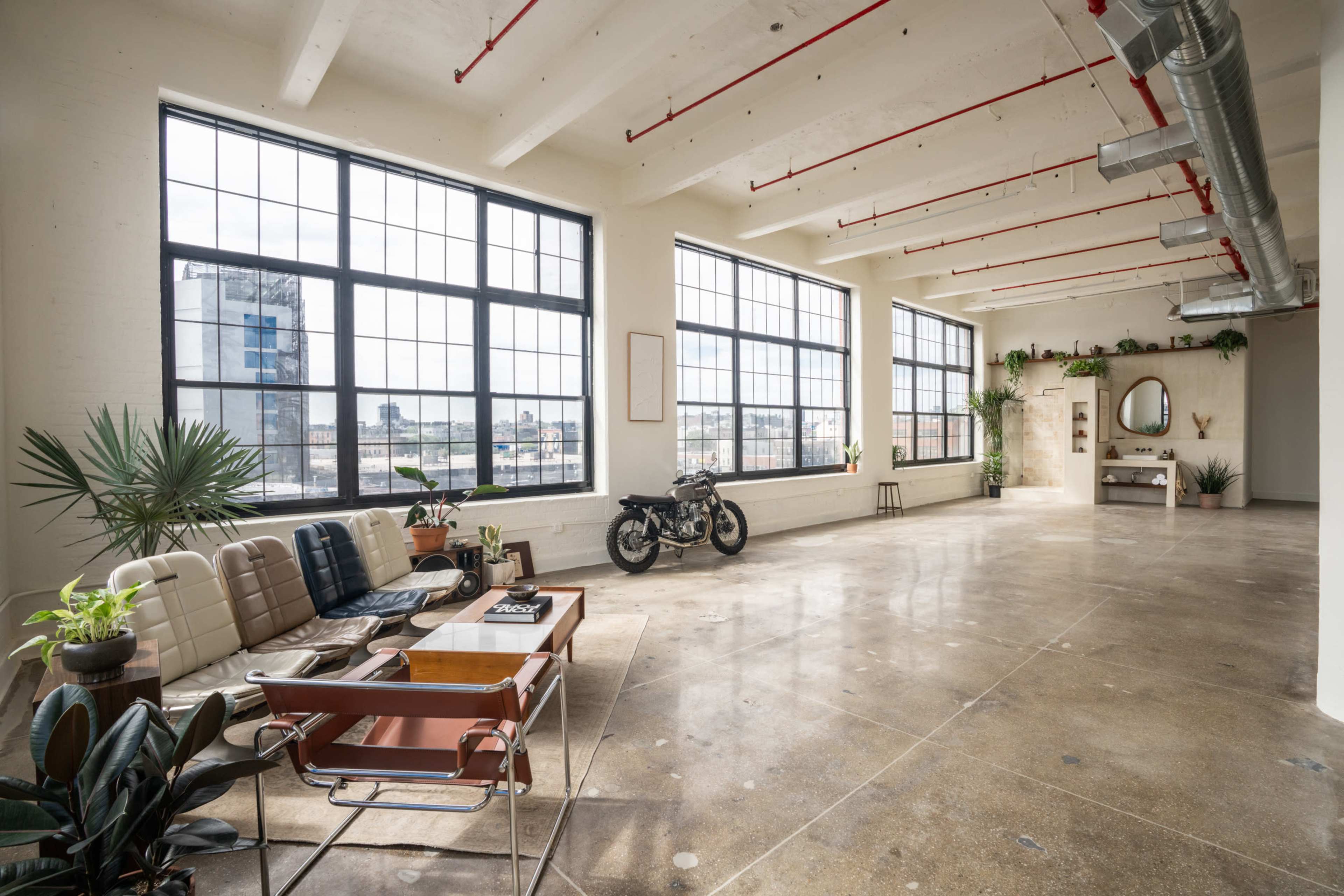 The image shows a spacious, well-lit industrial-style room with large windows, concrete floors, plants, a motorcycle, and minimalist furniture.