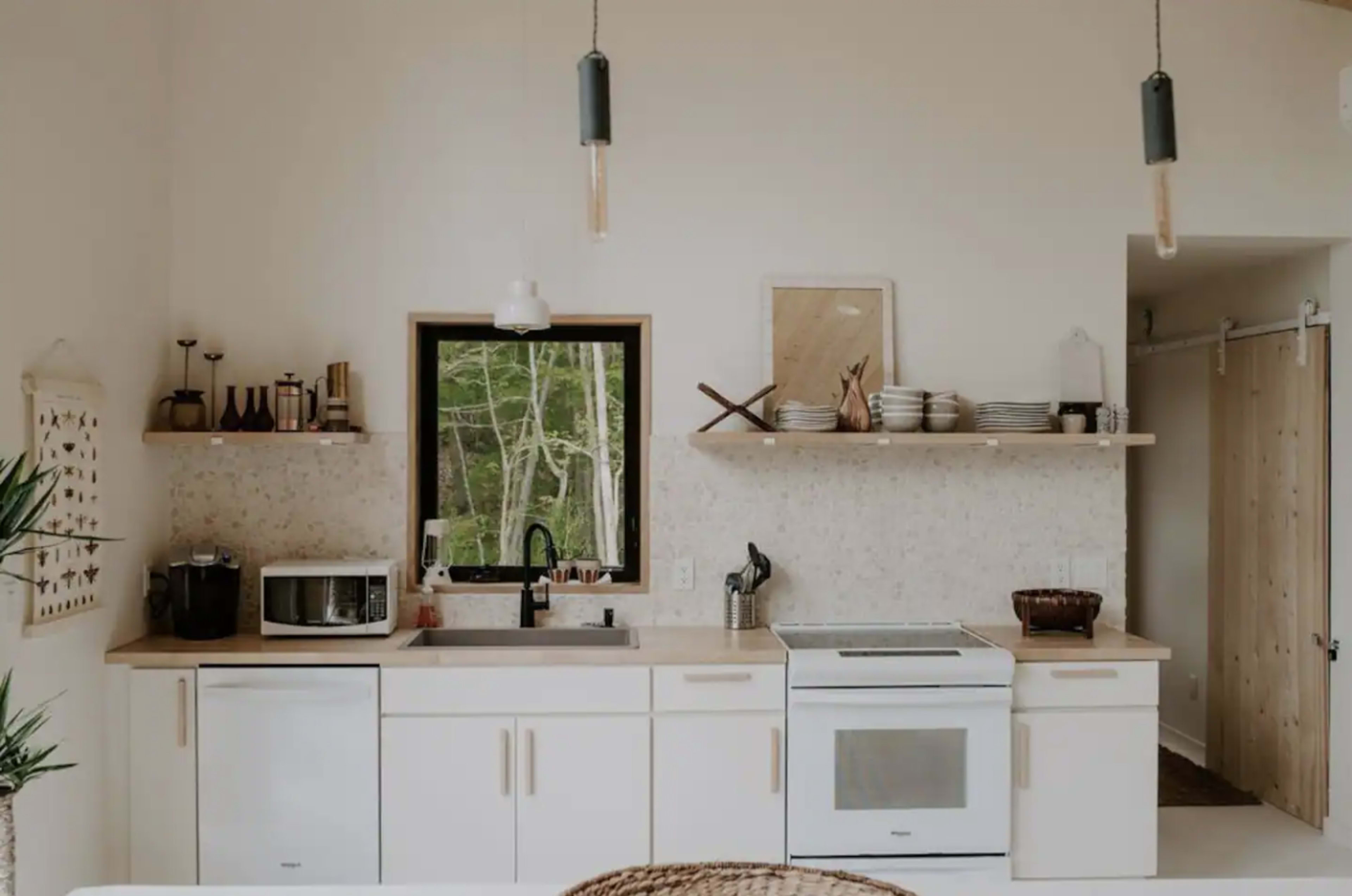 The image shows a modern kitchen with white cabinetry, a countertop, and an array of appliances, including a microwave and an oven, against a backdrop of natural light and greenery visible through a window.