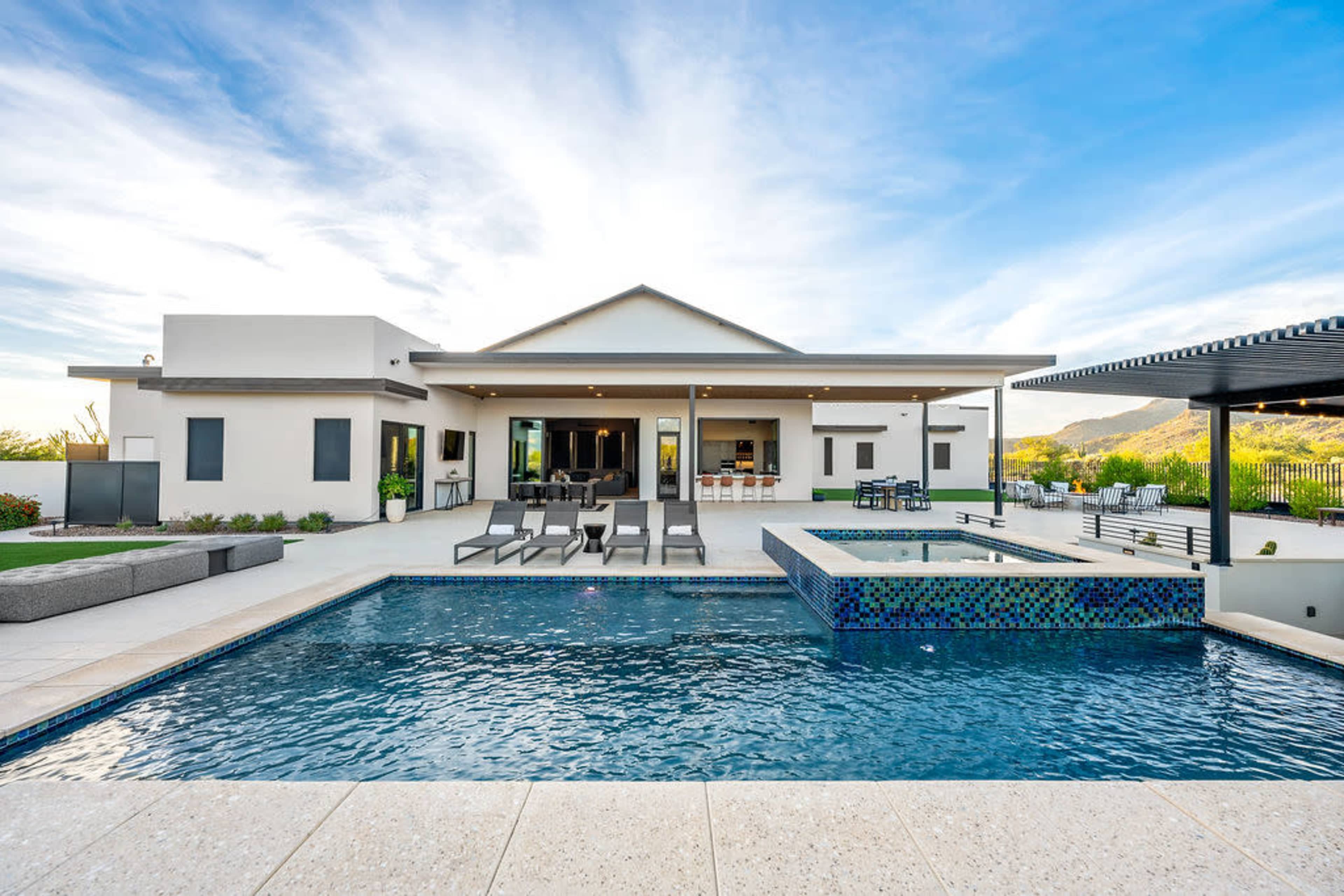 The image shows a modern house with a swimming pool and lounge area, surrounded by landscaped grounds and mountains in the background.
