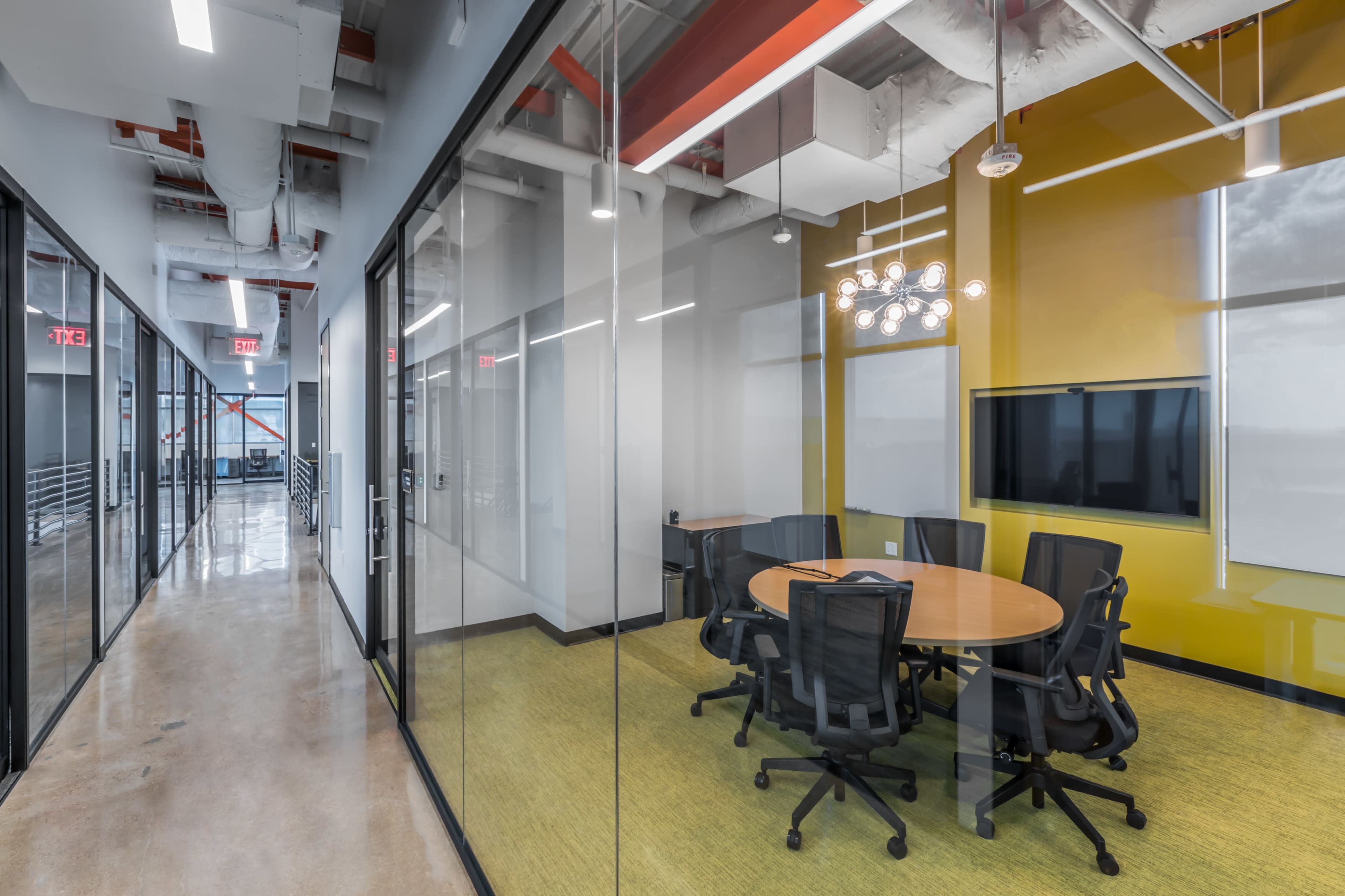 The image shows a modern office space with glass-walled conference rooms, a round table surrounded by black chairs, and bright yellow walls.