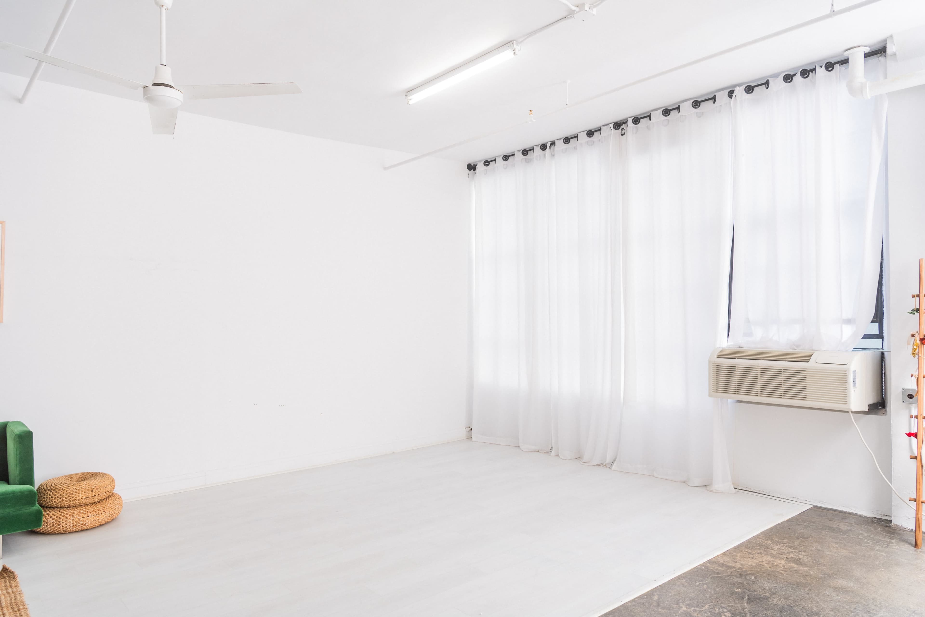 A spacious, minimally furnished room with white walls, a fan, large curtains, and an air conditioning unit.